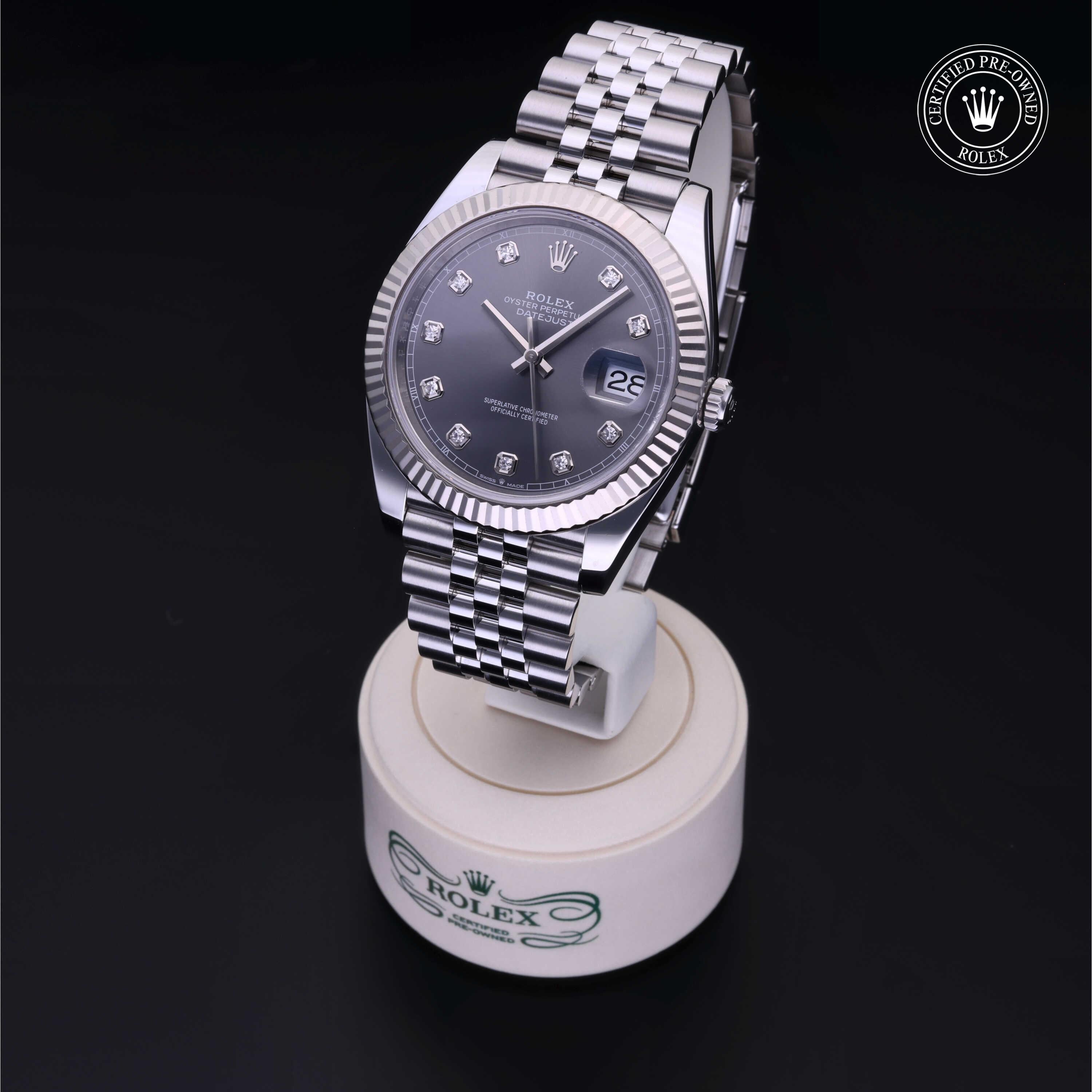 Rolex watch with a silver metal bracelet and diamond hour markers displayed on a white stand against a black background, featuring a certified pre-owned logo.