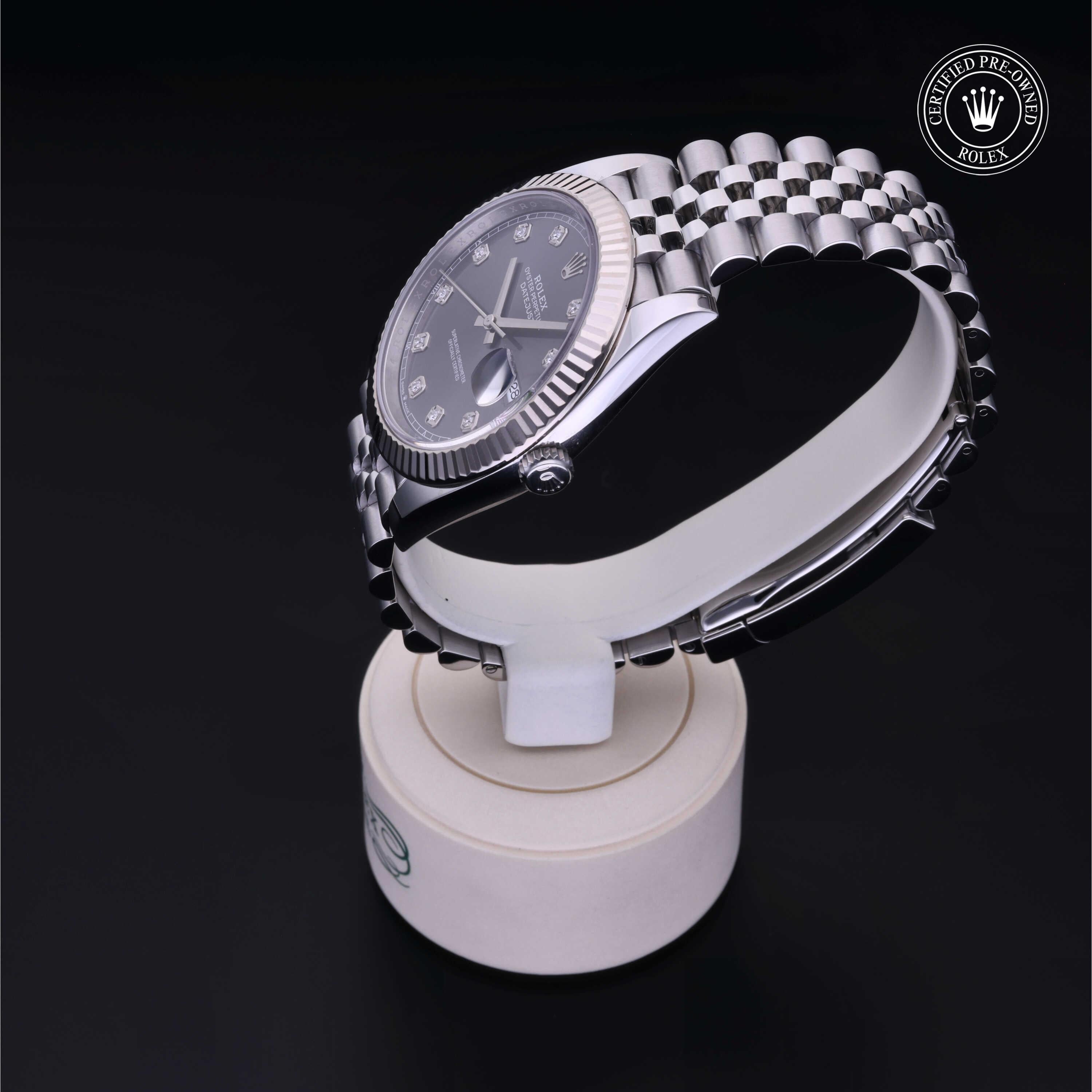 Silver luxury Rolex watch with a black dial and diamond hour markers displayed on a white stand against a black background, featuring a certified pre-owned Rolex emblem.