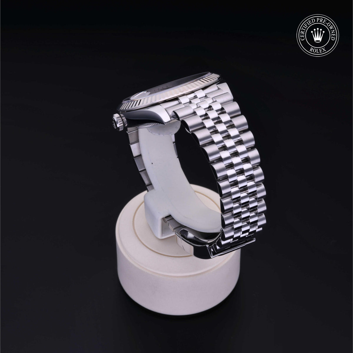 Side view of a luxury Rolex watch with a polished silver metal bracelet displayed on a white stand against a black background, featuring a certified pre-owned emblem.