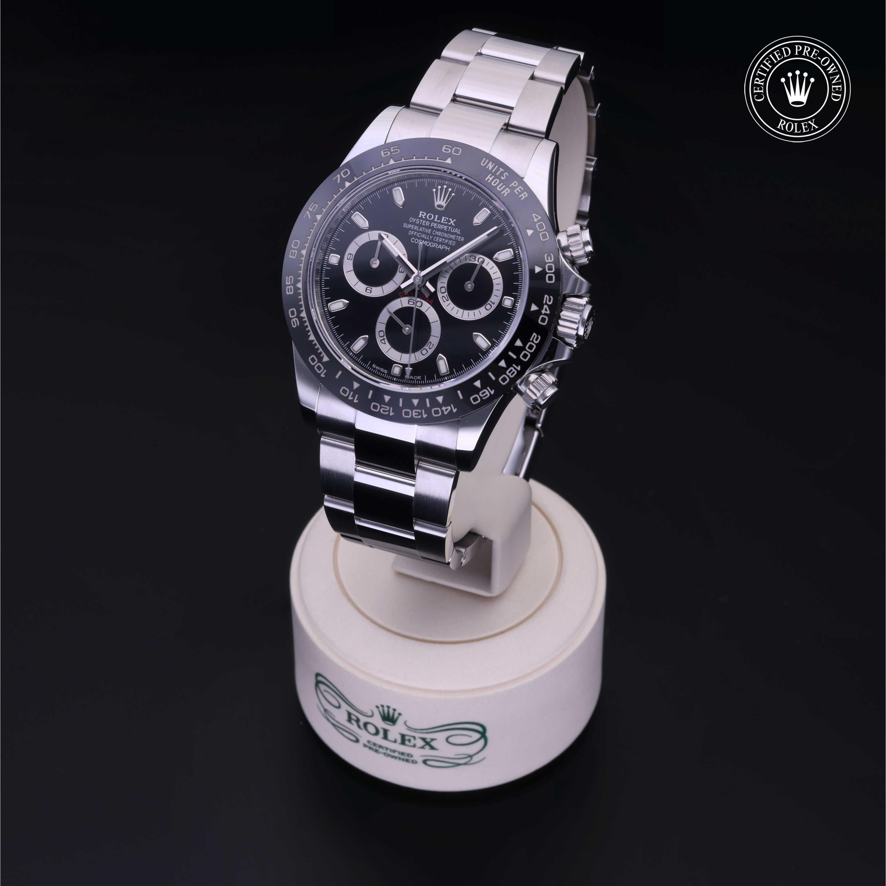 A stainless steel Rolex watch with a black dial and three subdials is displayed on a white stand with the Rolex logo, alongside a certified pre-owned Rolex badge.