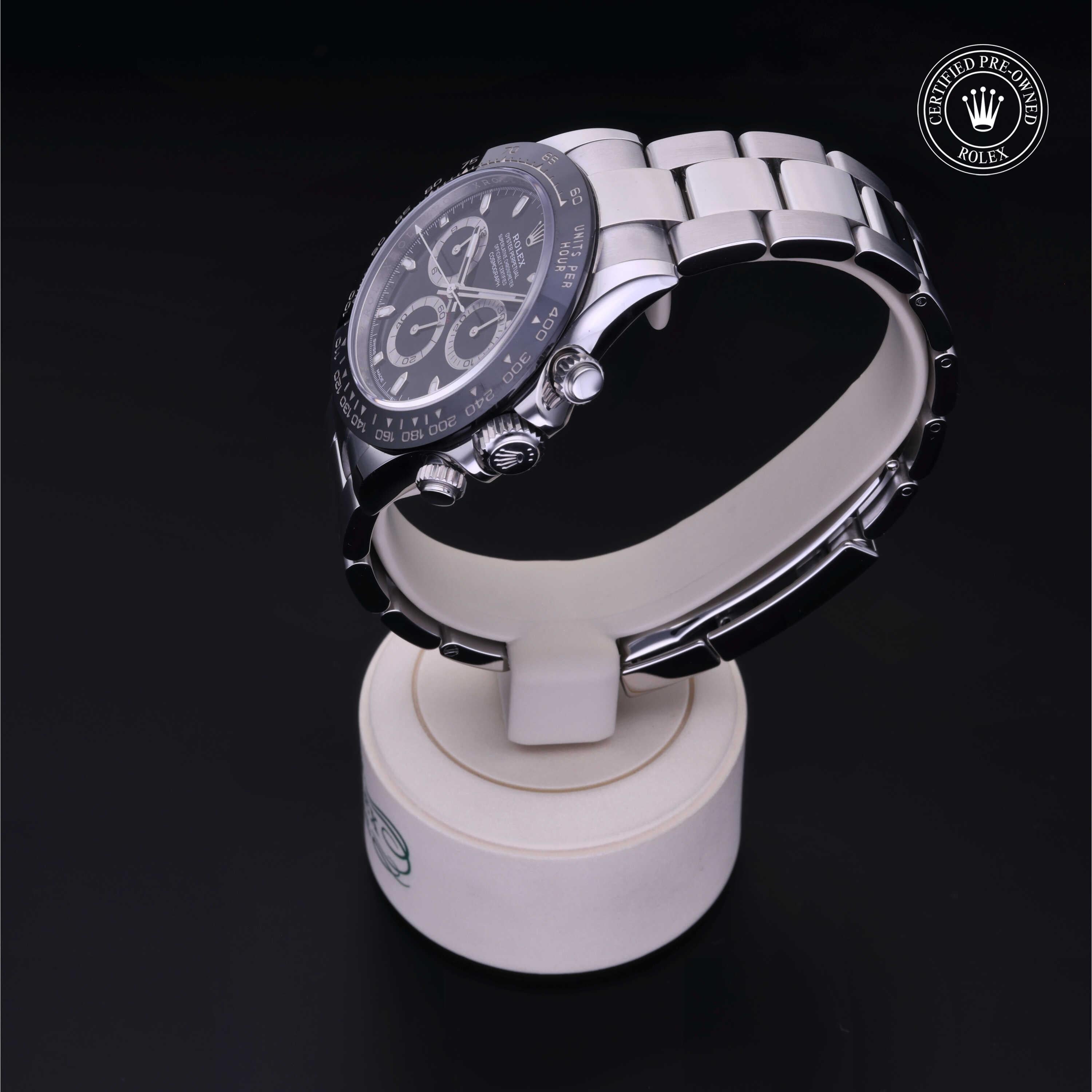 A luxury stainless steel Rolex Daytona watch with a black dial and chronograph features displayed on a white stand against a dark background, featuring Rolex Certified Pre-Owned logo.