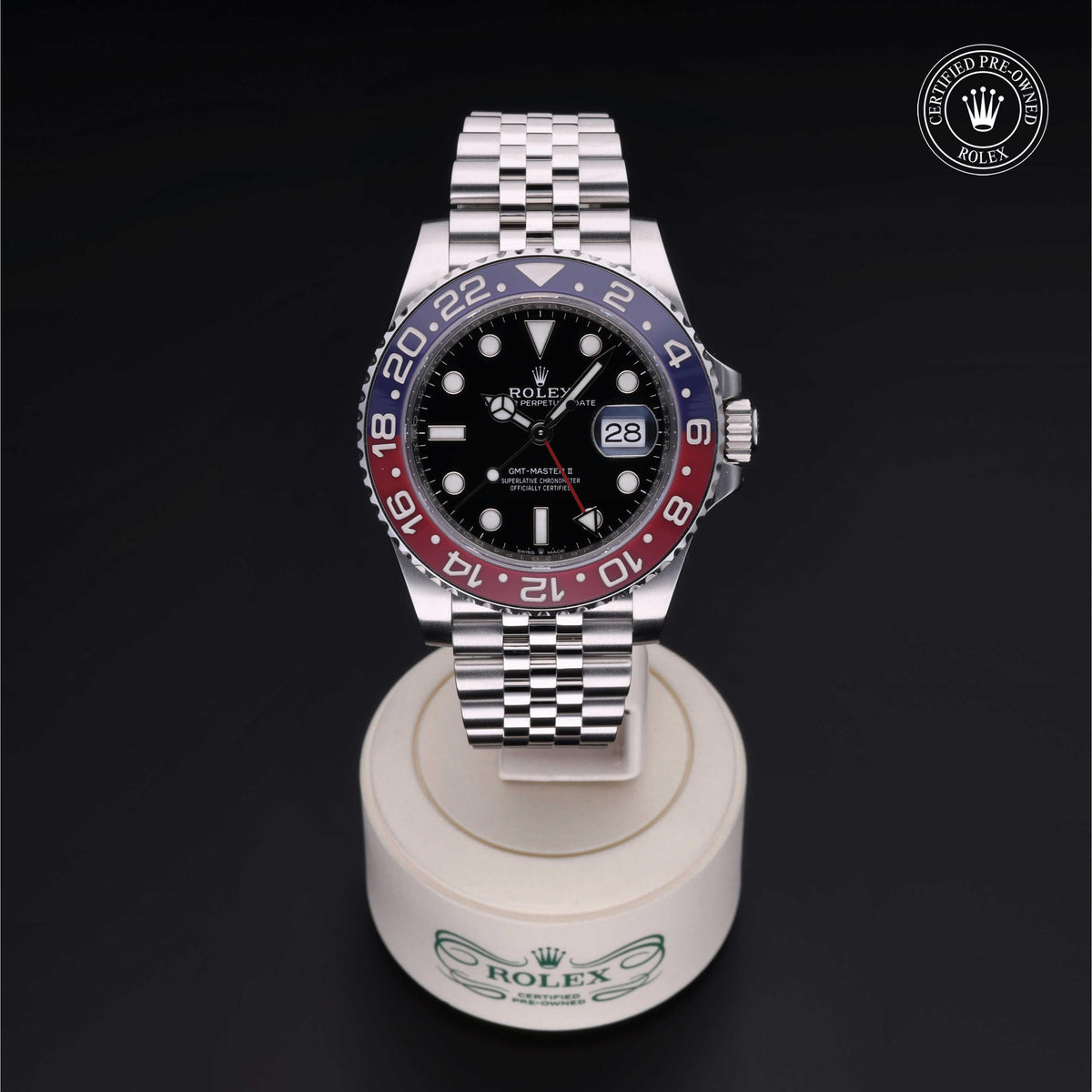 Rolex luxury watch with a red and blue bezel and black dial, displayed on a branded stand with a certified pre-owned Rolex seal in the corner.