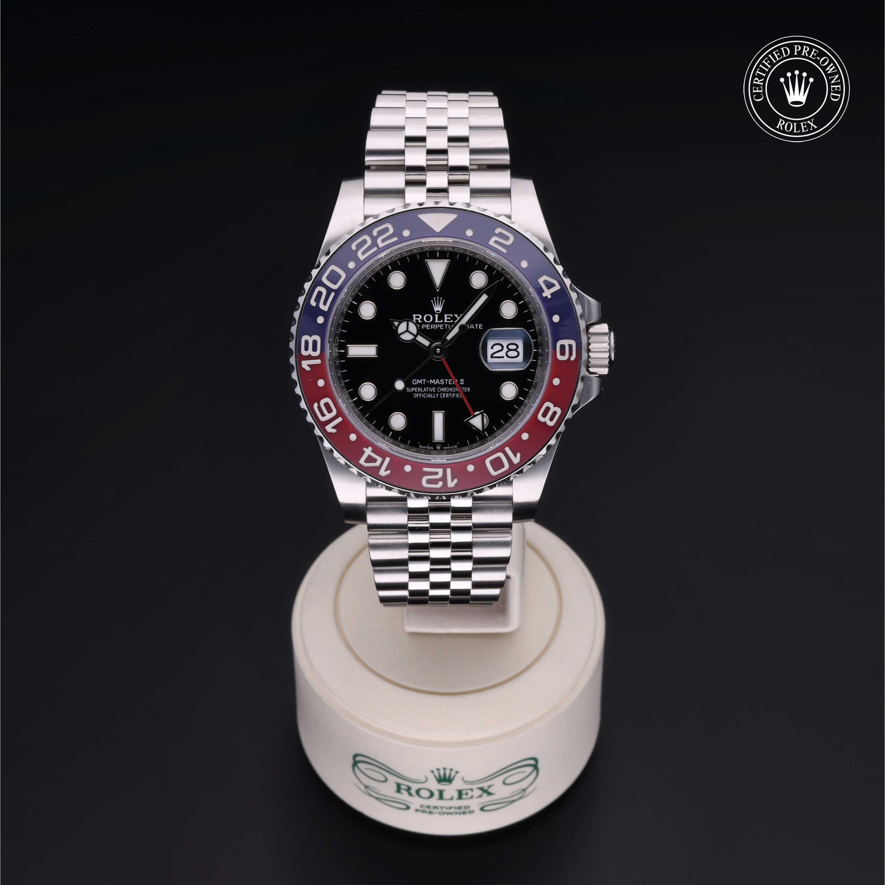 Rolex luxury watch with a red and blue bezel and black dial, displayed on a branded stand with a certified pre-owned Rolex seal in the corner.