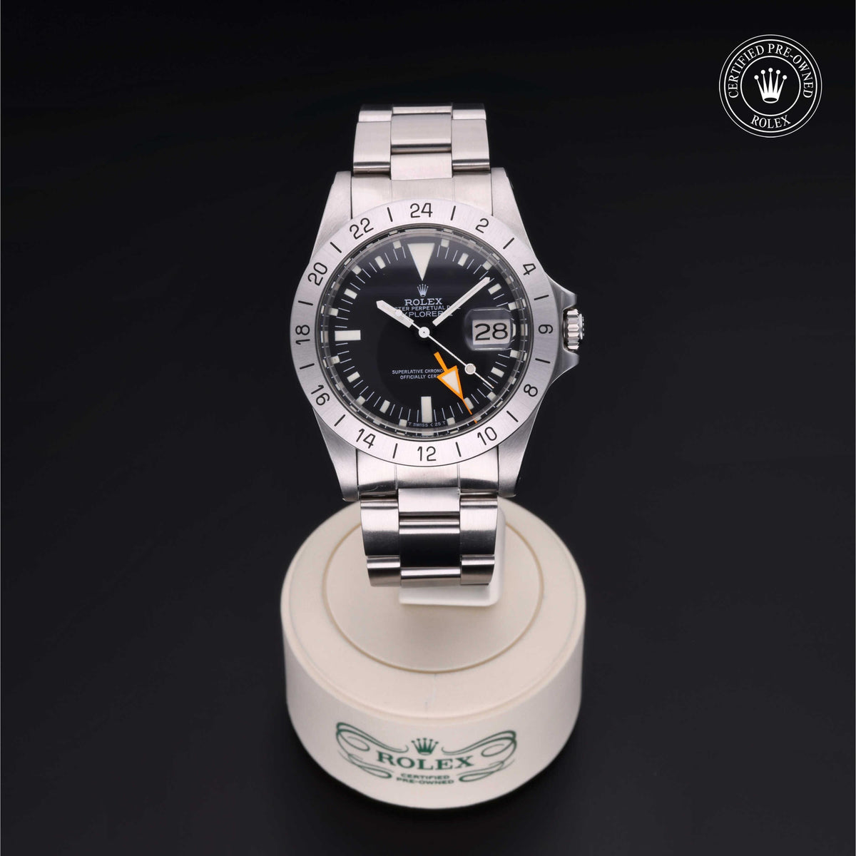 Stainless steel Rolex watch with a black dial and orange GMT hand displayed on a white stand featuring the Rolex logo, with a certified pre-owned Rolex emblem above.