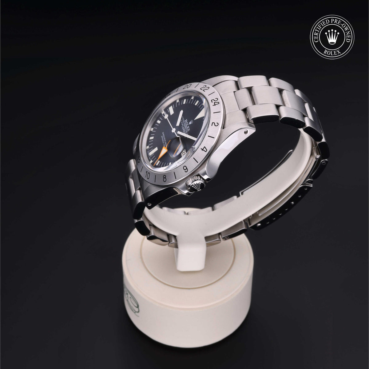 Stainless steel Rolex watch with a black dial and orange hand displayed on a white stand against a black background, featuring a certified pre-owned Rolex logo.