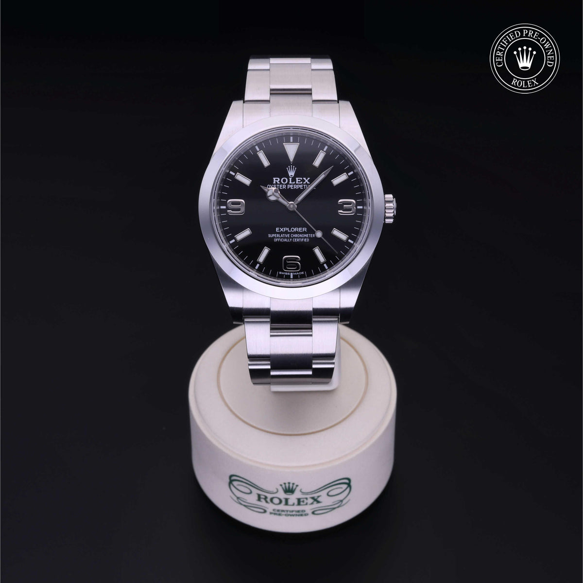 A stainless steel Rolex Explorer with a black dial and silver hour markers displayed on a round white stand with Rolex branding, accompanied by a certified pre-owned Rolex logo.