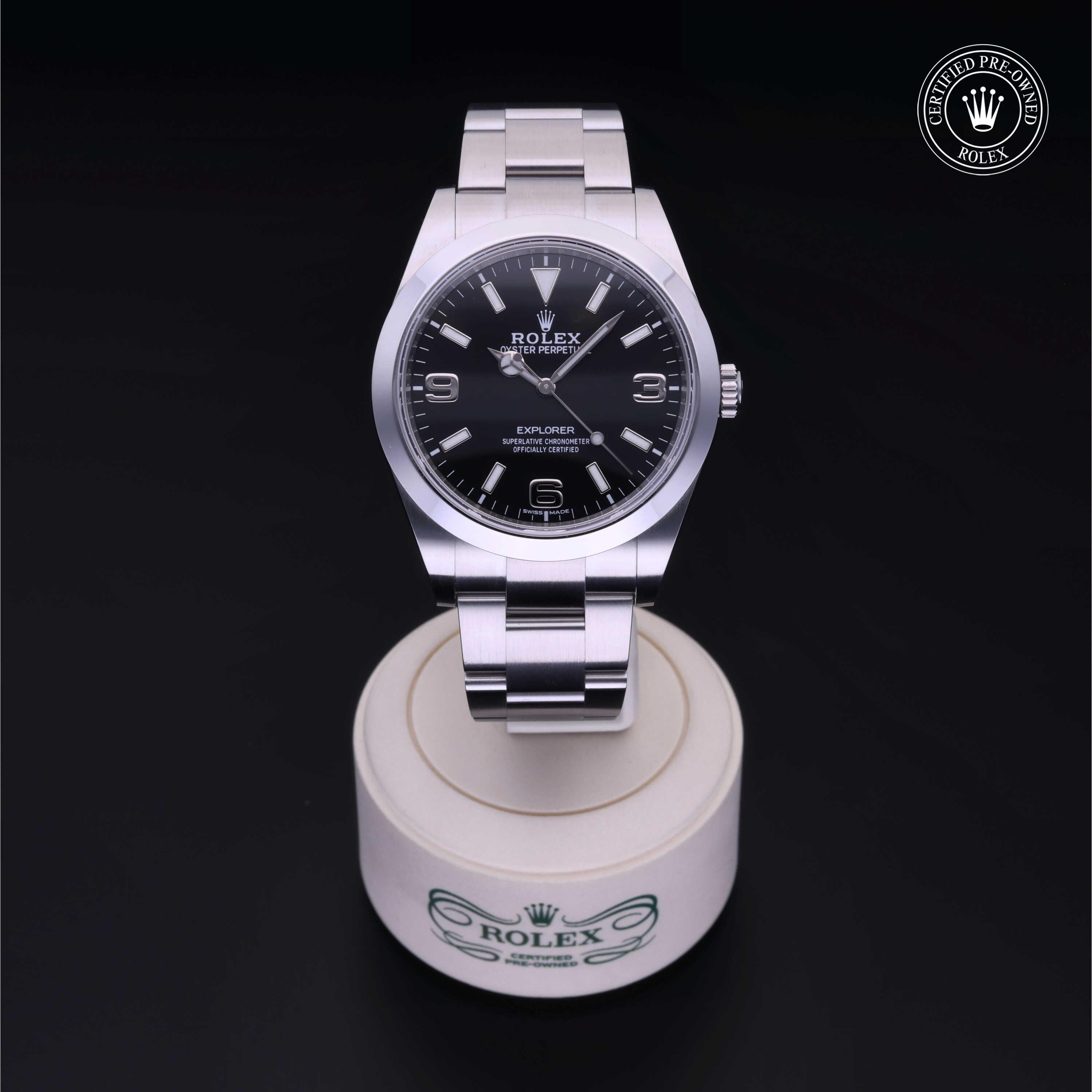 A stainless steel Rolex Explorer with a black dial and silver hour markers displayed on a round white stand with Rolex branding, accompanied by a certified pre-owned Rolex logo.