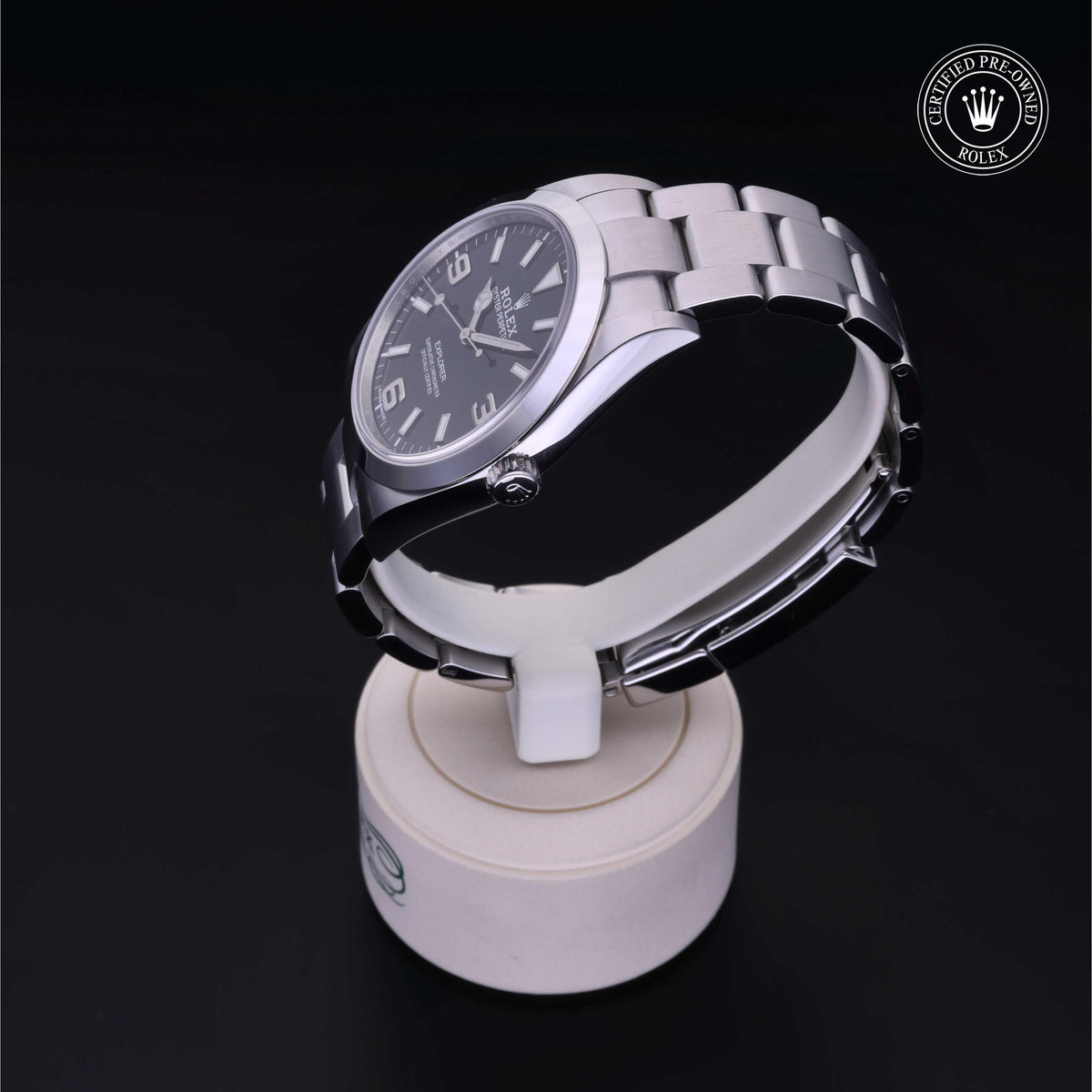 Silver stainless steel Rolex watch with a black dial featuring numeric hour markers, displayed on a white stand against a dark background with a certified pre-owned Rolex emblem.