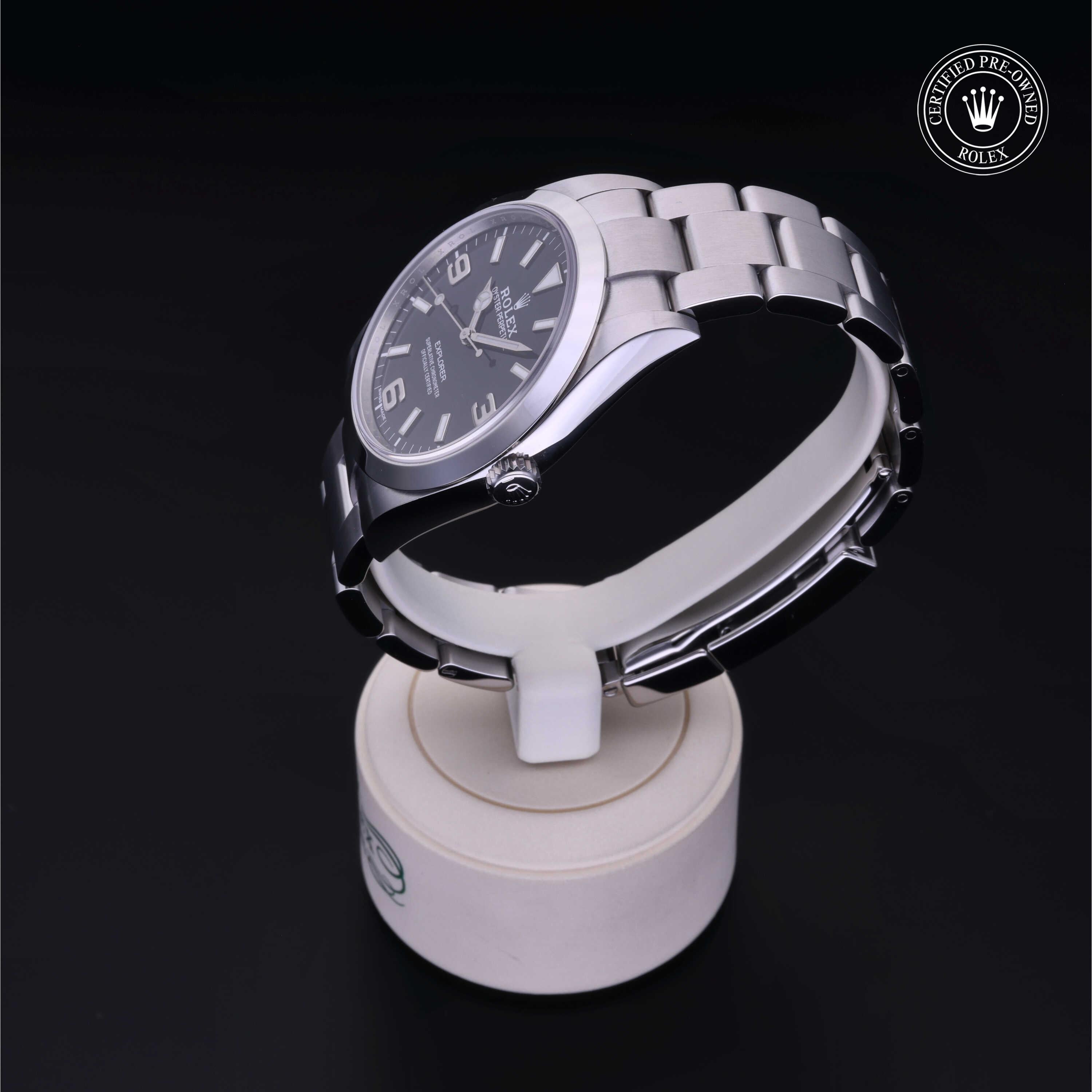 Silver stainless steel Rolex watch with a black dial featuring numeric hour markers, displayed on a white stand against a dark background with a certified pre-owned Rolex emblem.