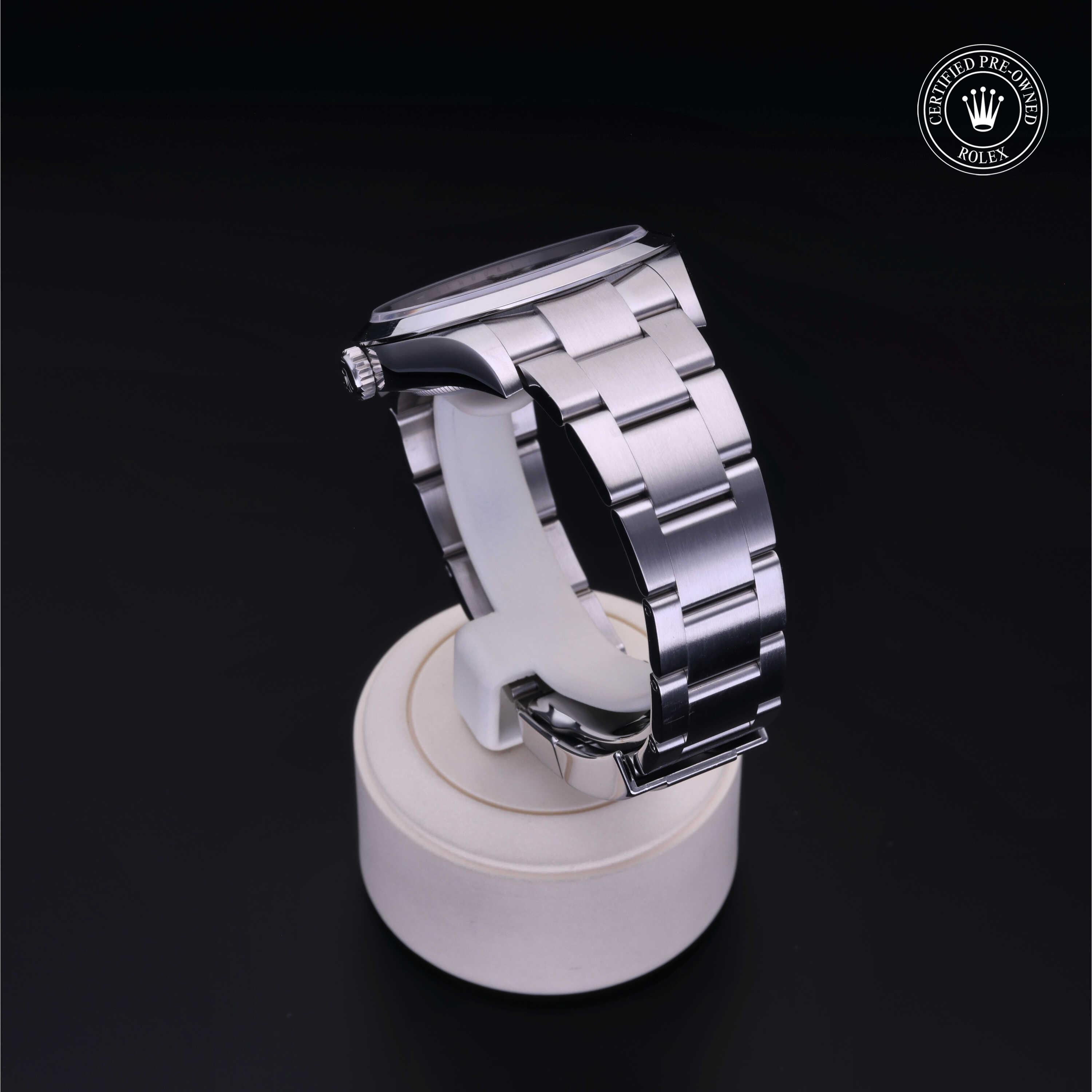 Silver-tone luxury watch with a brushed metal bracelet displayed on a white stand against a dark background, featuring a certified pre-owned Rolex emblem.