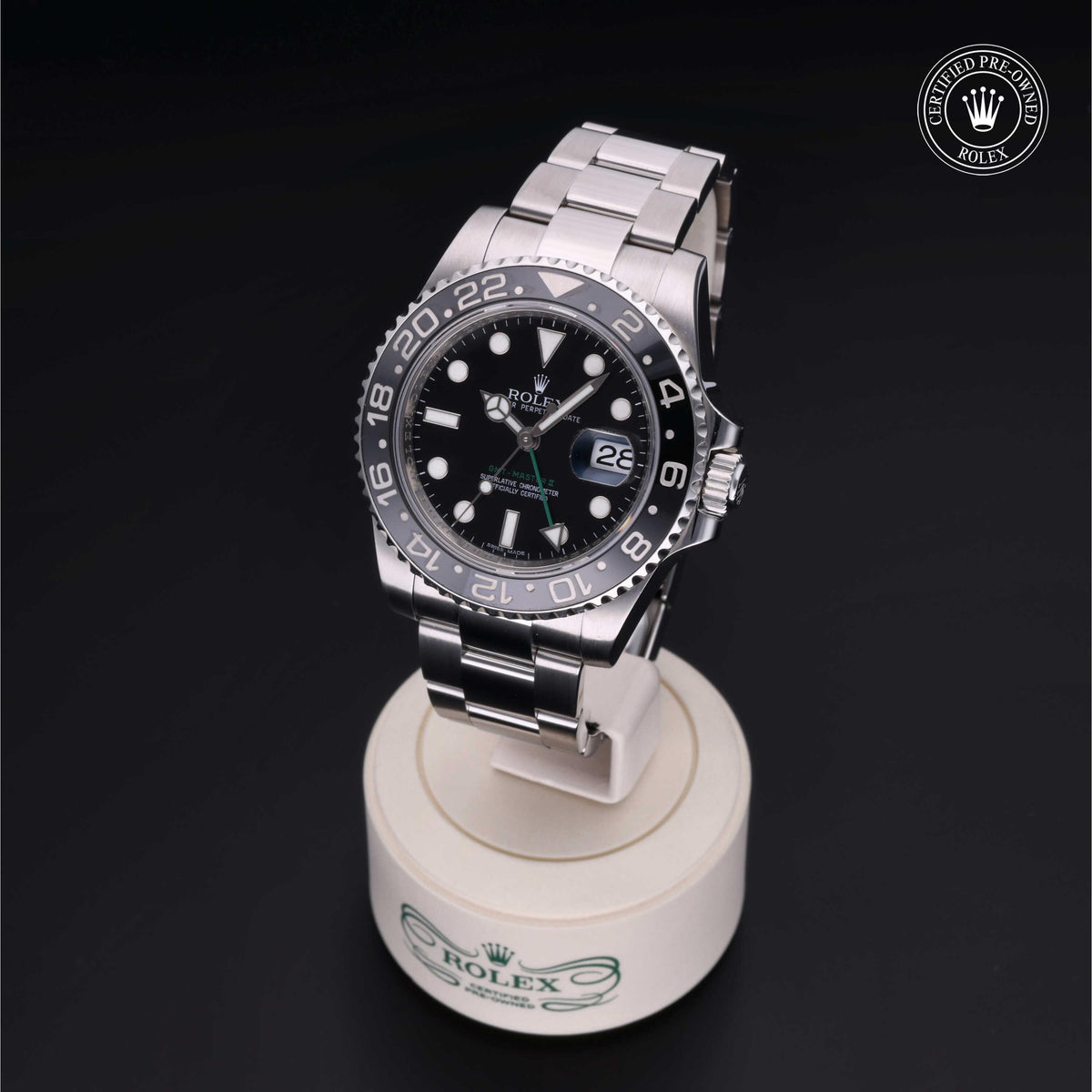 Rolex luxury watch with black dial and silver bezel displayed on a white stand marked with the Rolex logo against a black background, certified pre-owned badge visible.