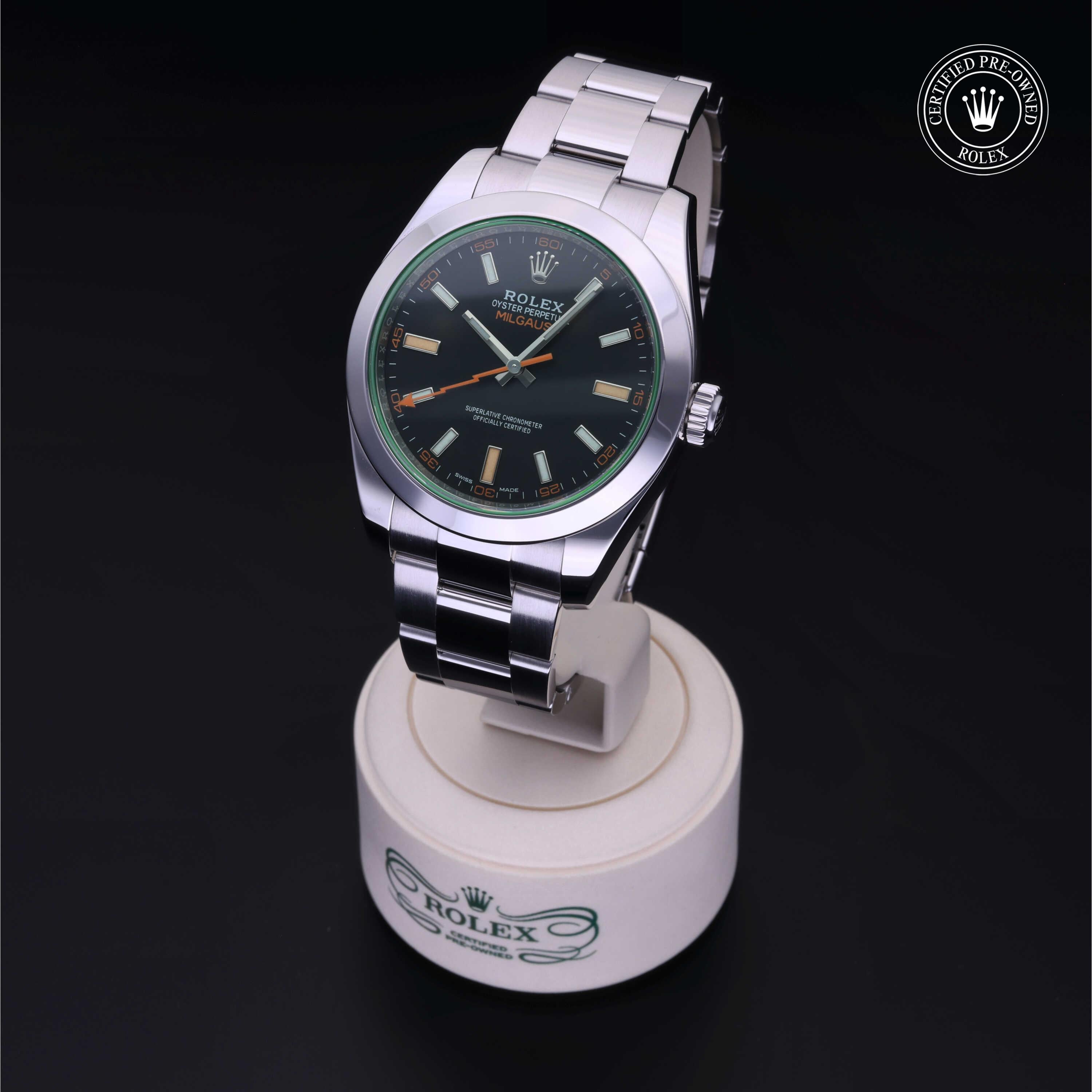 Stainless steel Rolex Milgauss watch with black dial, orange lightning bolt second hand, and green sapphire crystal, displayed on a white stand with Rolex logo and certified pre-owned badge.