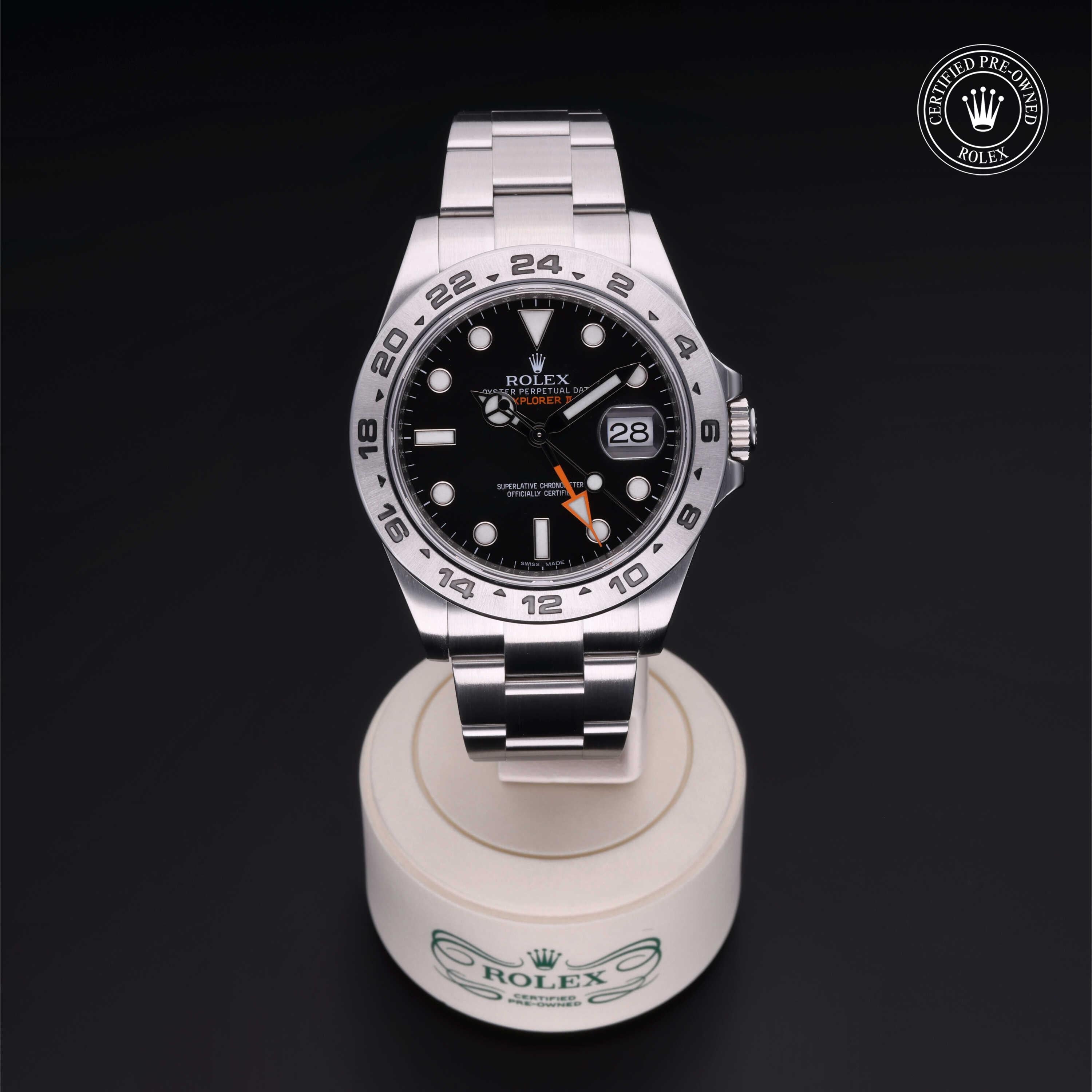 Stainless steel Rolex Explorer wristwatch with black dial, orange 24-hour hand, date window, and Oyster bracelet displayed on a branded white stand against a black background.