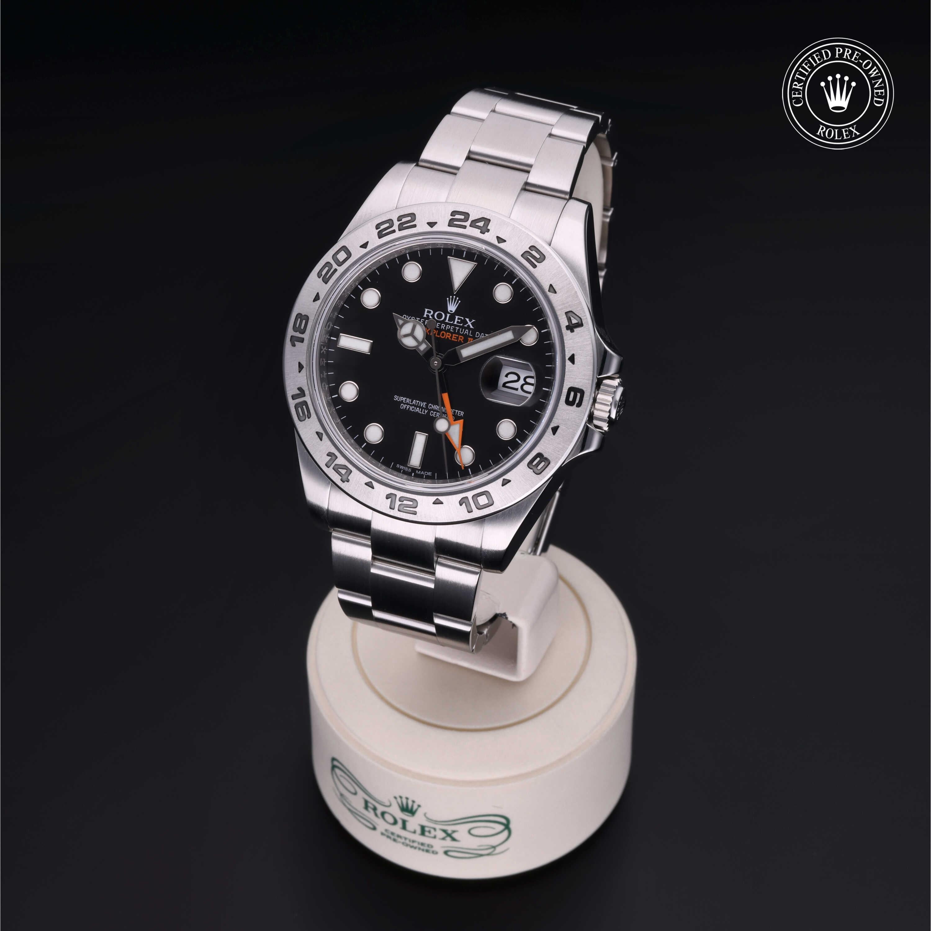 Rolex stainless steel watch with black dial, orange GMT hand, and silver 24-hour bezel, displayed on a branded stand with certified pre-owned Rolex logo.
