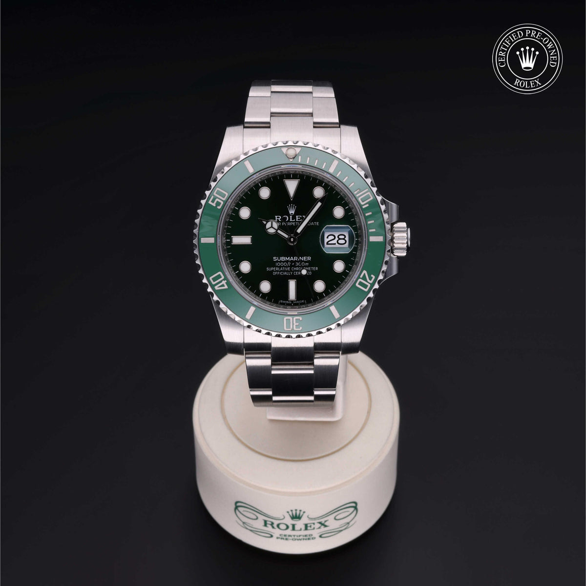 Silver Rolex Submariner watch with green bezel and black dial displayed on a white stand against a black background, featuring a certified pre-owned Rolex logo badge.