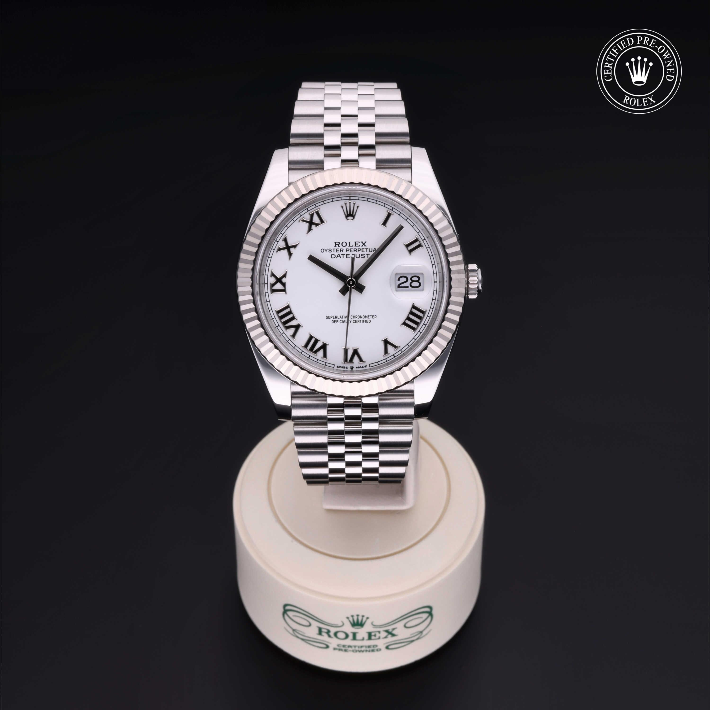 Rolex wristwatch with white dial, black Roman numerals, and date display on a silver metal bracelet, displayed on a branded stand with luxury watch certification badge.