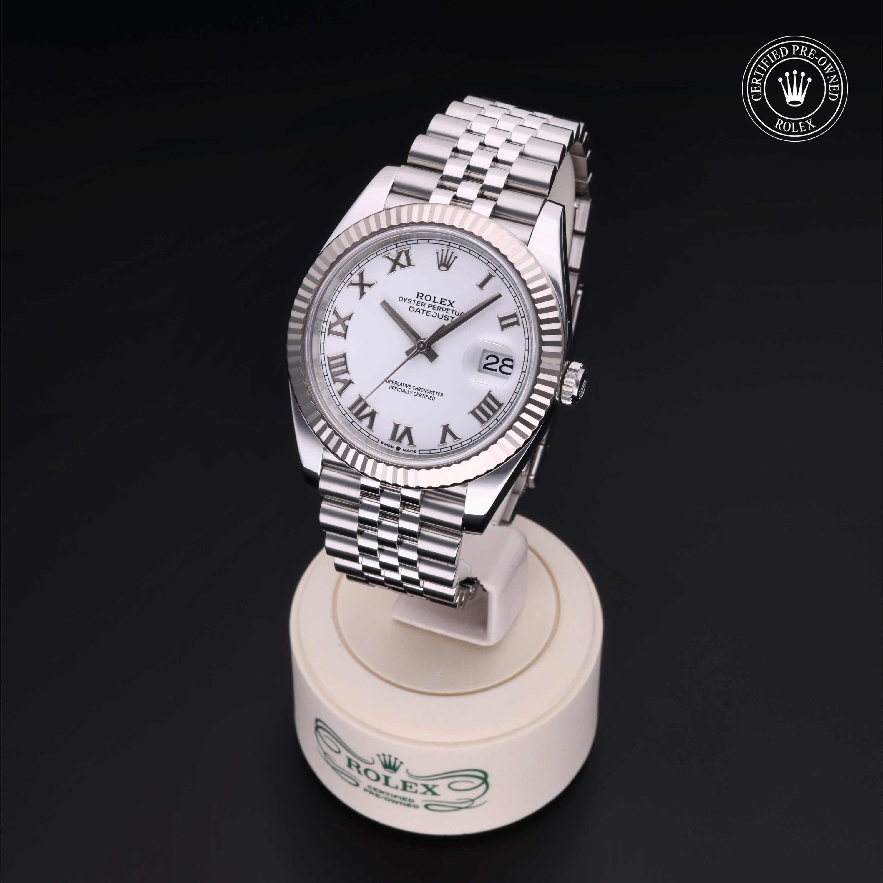 Rolex watch with white dial and roman numerals displayed on a stand with Rolex branding, featuring a stainless steel bracelet and date window, certified pre-owned badge.