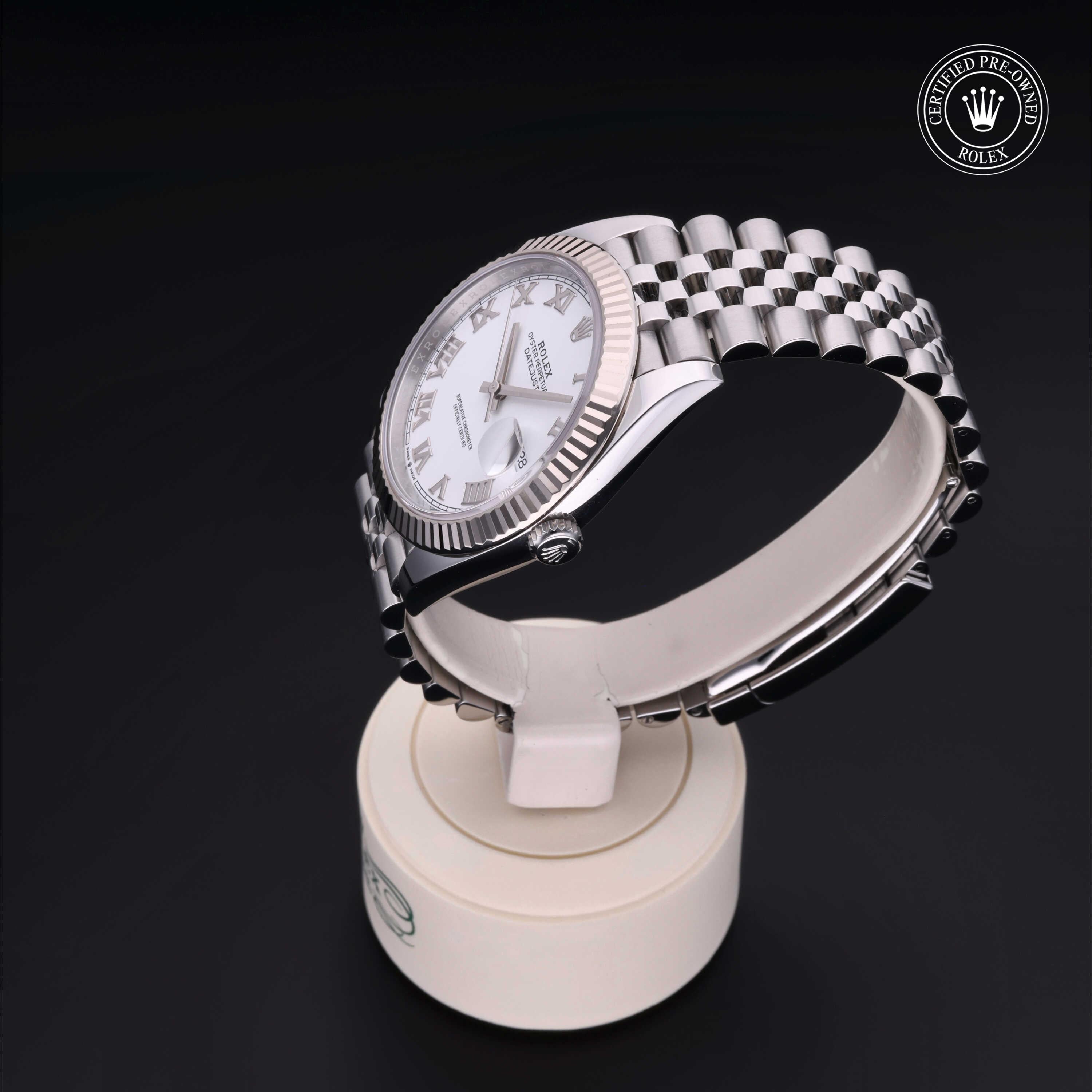 Silver Rolex watch with a white dial and Roman numerals displayed on a round white stand against a black background, showcasing luxury and craftsmanship.