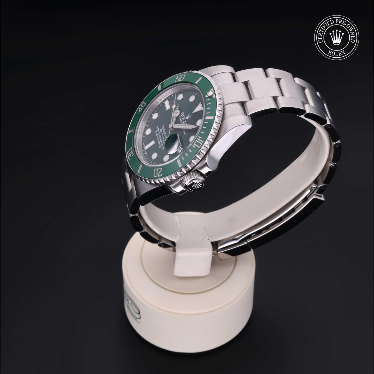 Silver Rolex watch with a green bezel displayed on a white stand, featuring a certified pre-owned Rolex emblem on a black background.