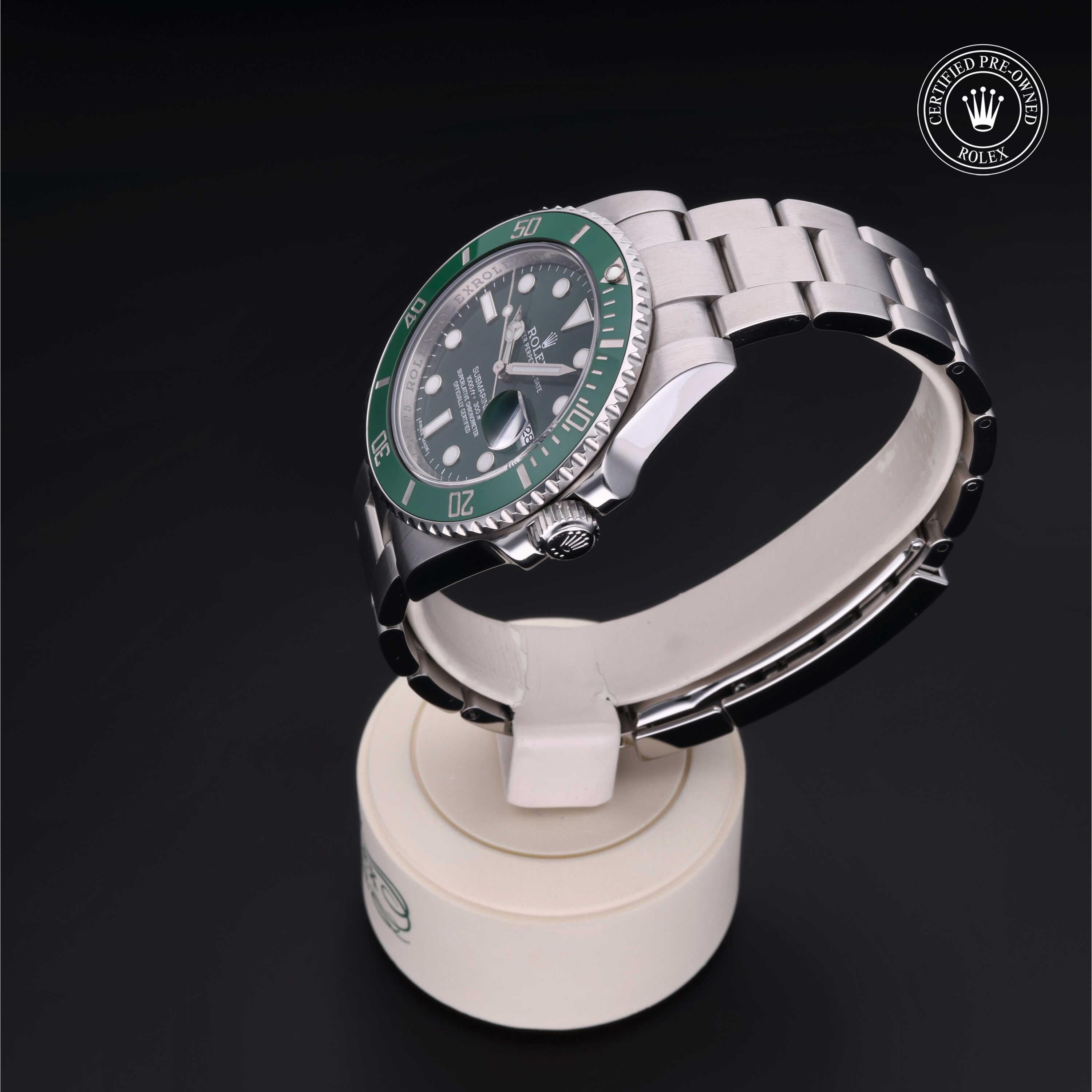 Silver Rolex watch with a green bezel displayed on a white stand, featuring a certified pre-owned Rolex emblem on a black background.