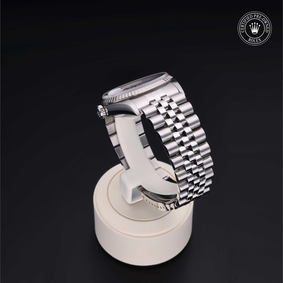 Silver luxury watch with a polished metal link bracelet displayed on a white stand against a black background, featuring a certified pre-owned Rolex logo in the corner.