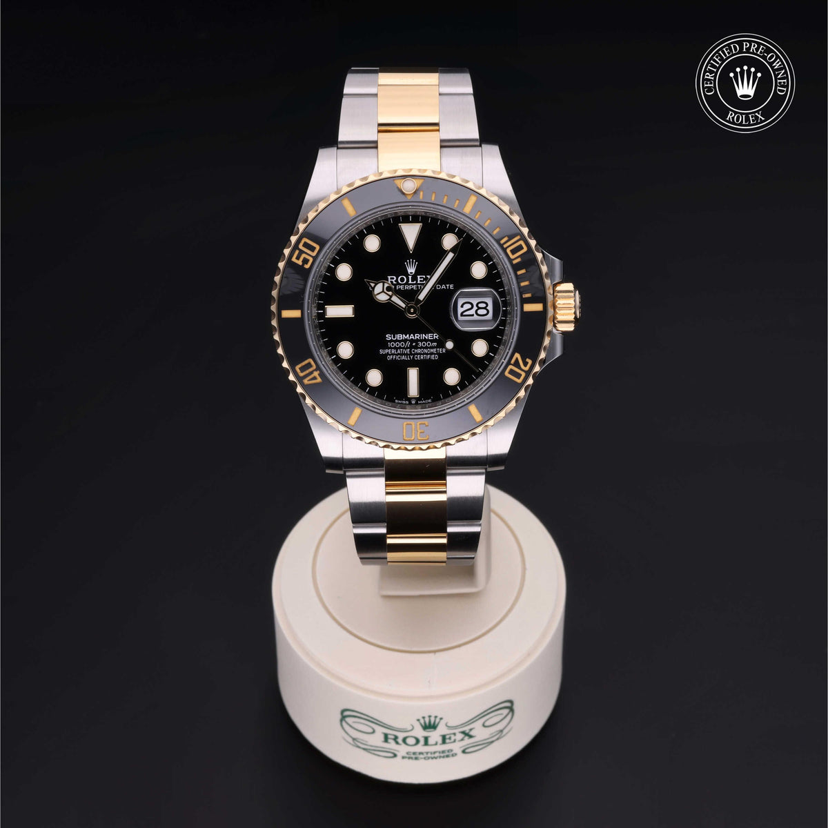 Rolex Submariner watch with black dial and gold and silver two-tone bracelet displayed on a white stand with Rolex logo, featuring certified pre-owned badge.