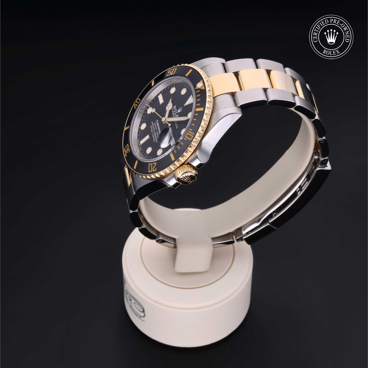 A two-tone stainless steel and gold Rolex luxury watch with a black dial and rotating bezel, displayed on a white stand against a black background, marked as certified pre-owned.
