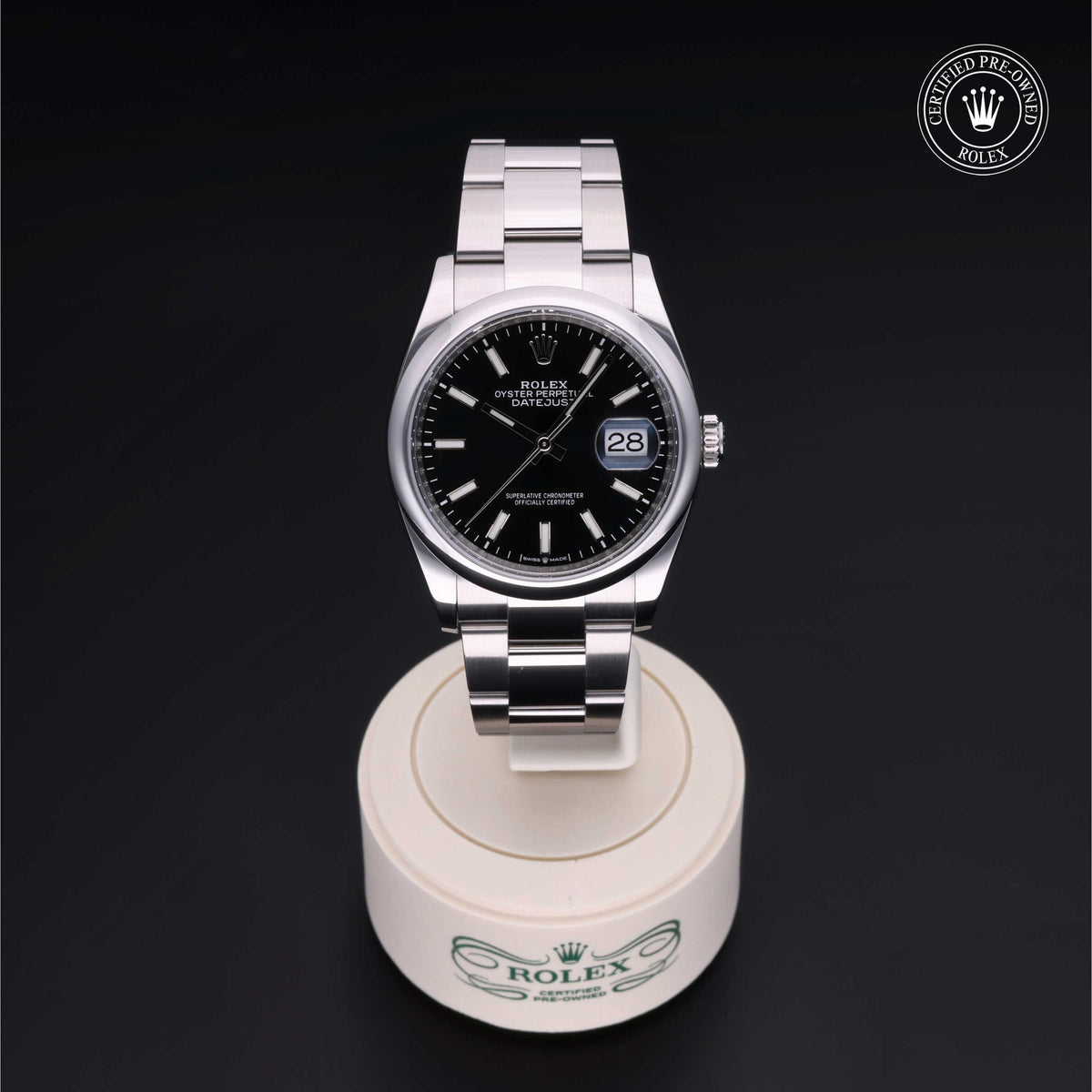 Rolex Datejust watch with black dial and stainless steel bracelet displayed on white stand with Rolex logo, featuring a date window and certified pre-owned emblem.