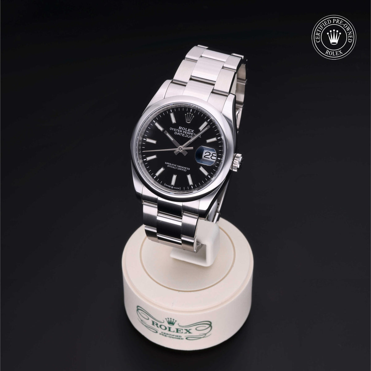 Stainless steel Rolex Datejust watch with black dial and date display on a cream-colored Rolex stand, featuring a luxury design certified pre-owned emblem in the corner.