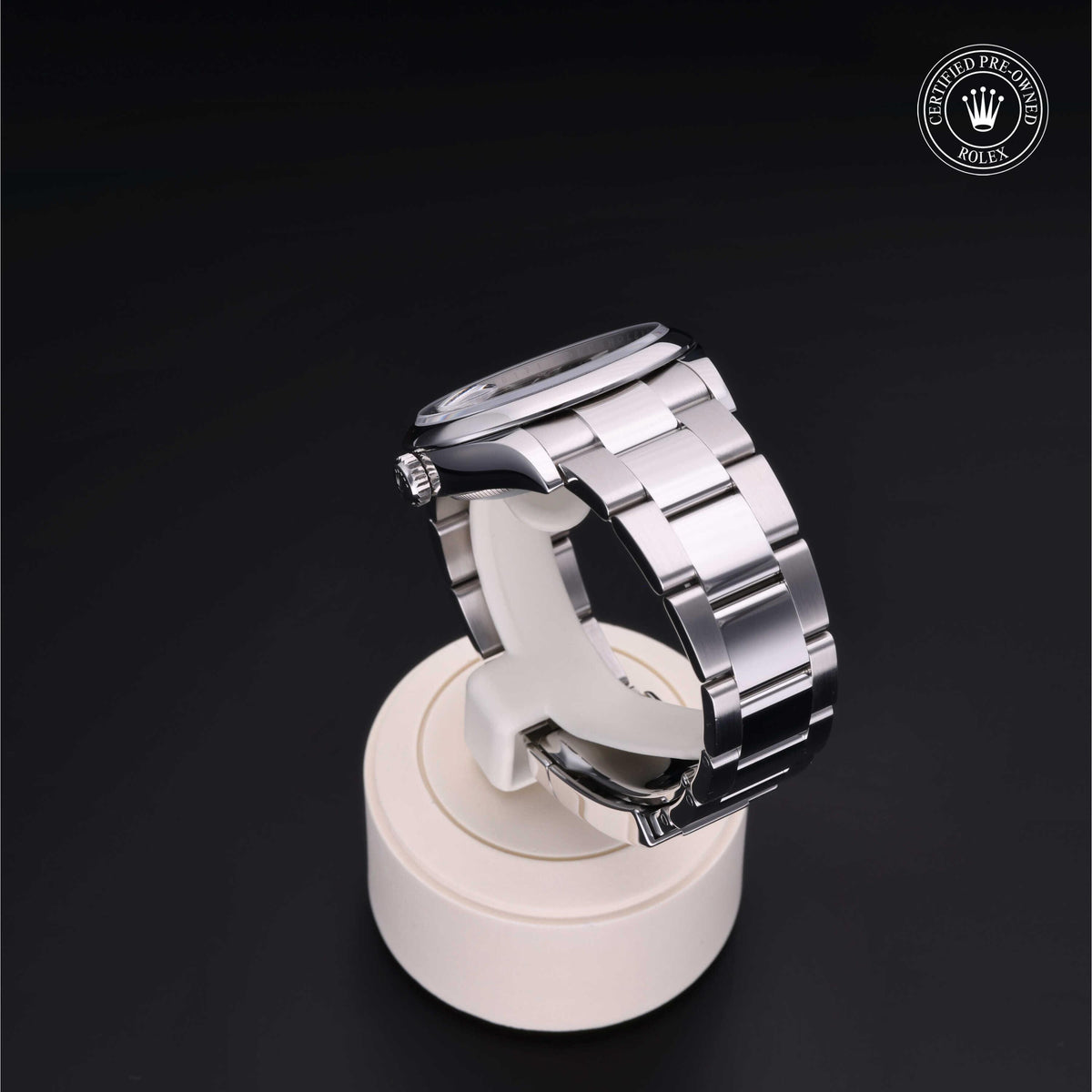 Silver Rolex watch with polished metal bracelet on a white display stand against a black background, featuring a certified pre-owned Rolex logo.