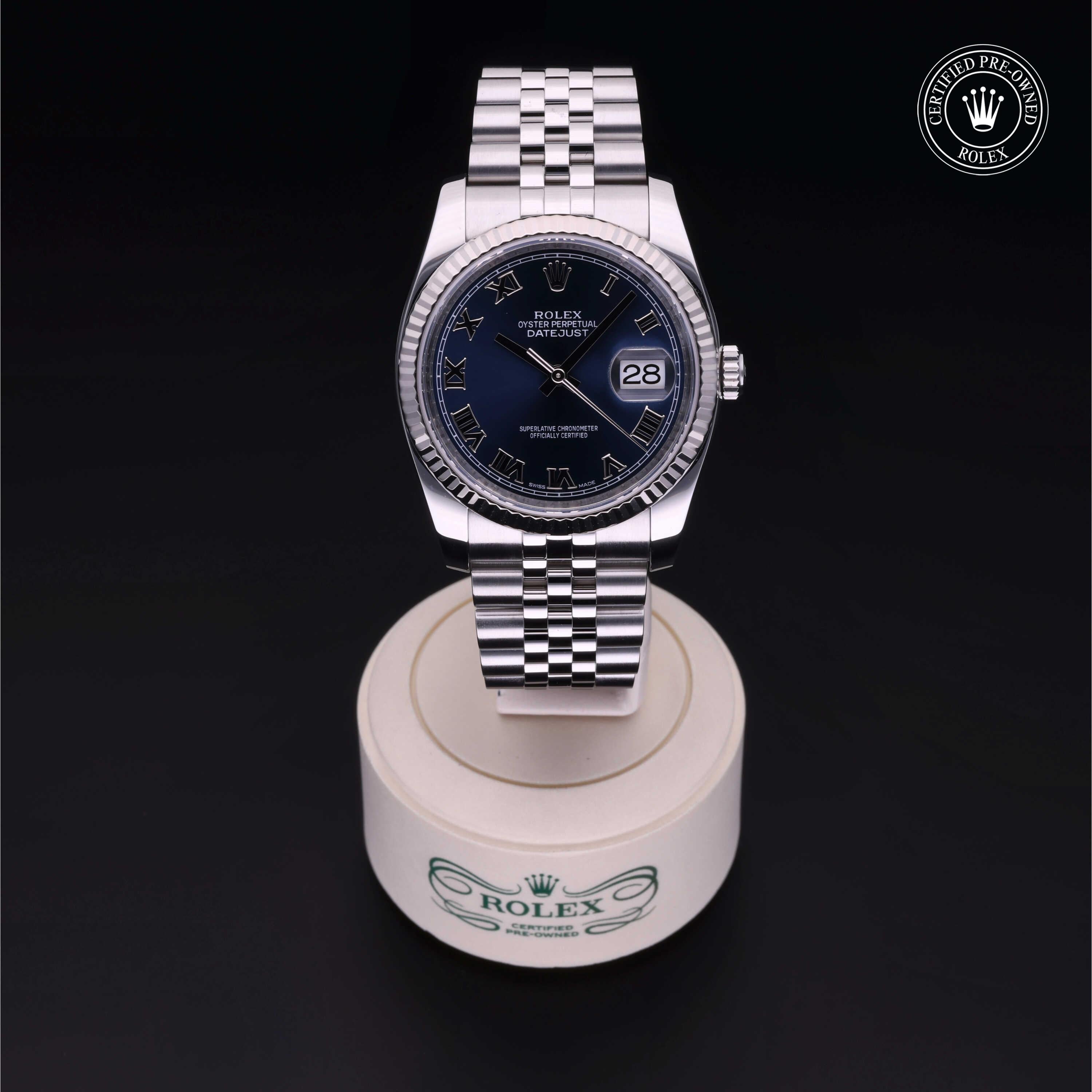Rolex Datejust wristwatch with a silver metal band and dark blue dial featuring Roman numerals, displayed on a round white holder with Rolex branding against a black background.