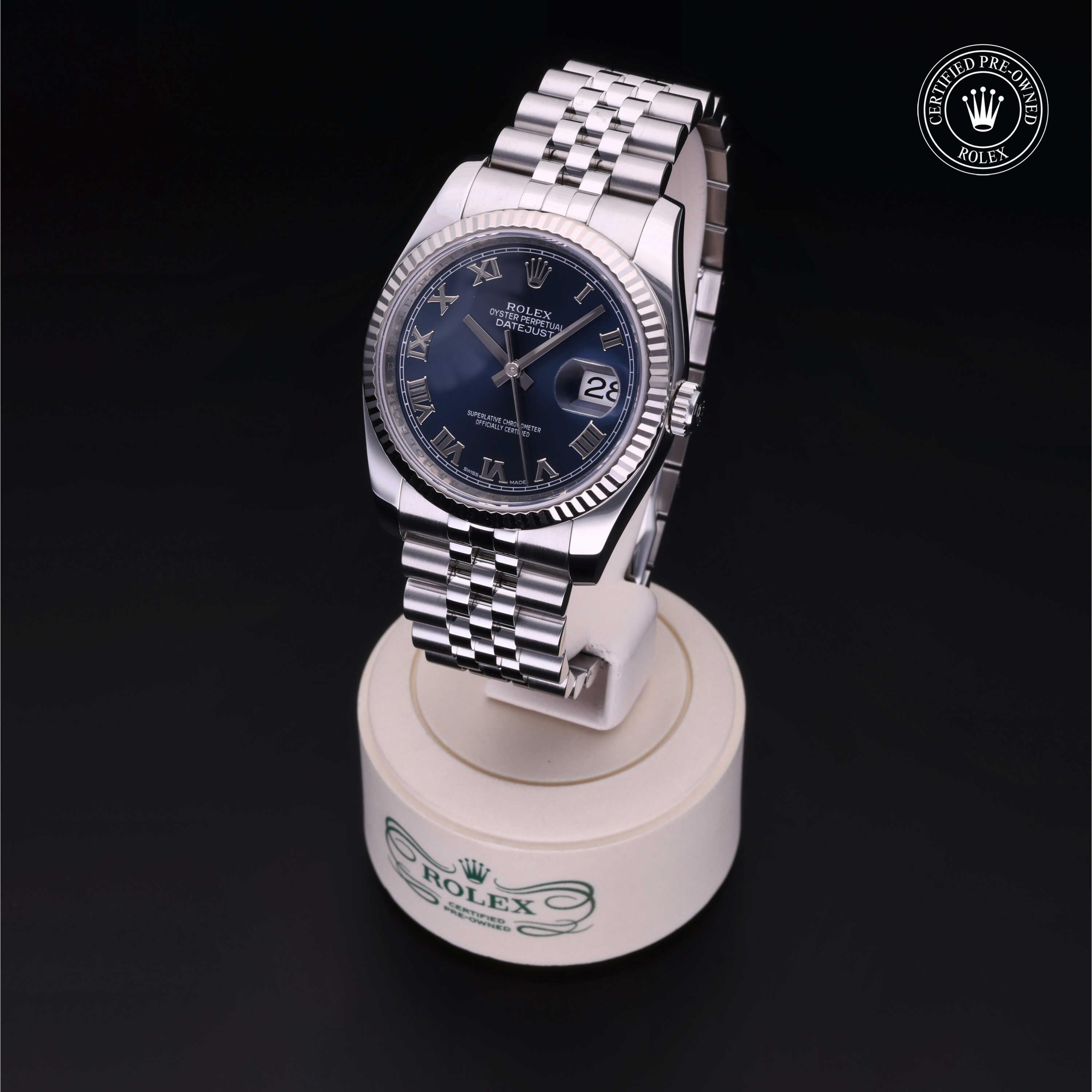 Rolex Datejust watch with a blue dial and Roman numerals on a stainless steel Jubilee bracelet, displayed on a branded stand against a black background.