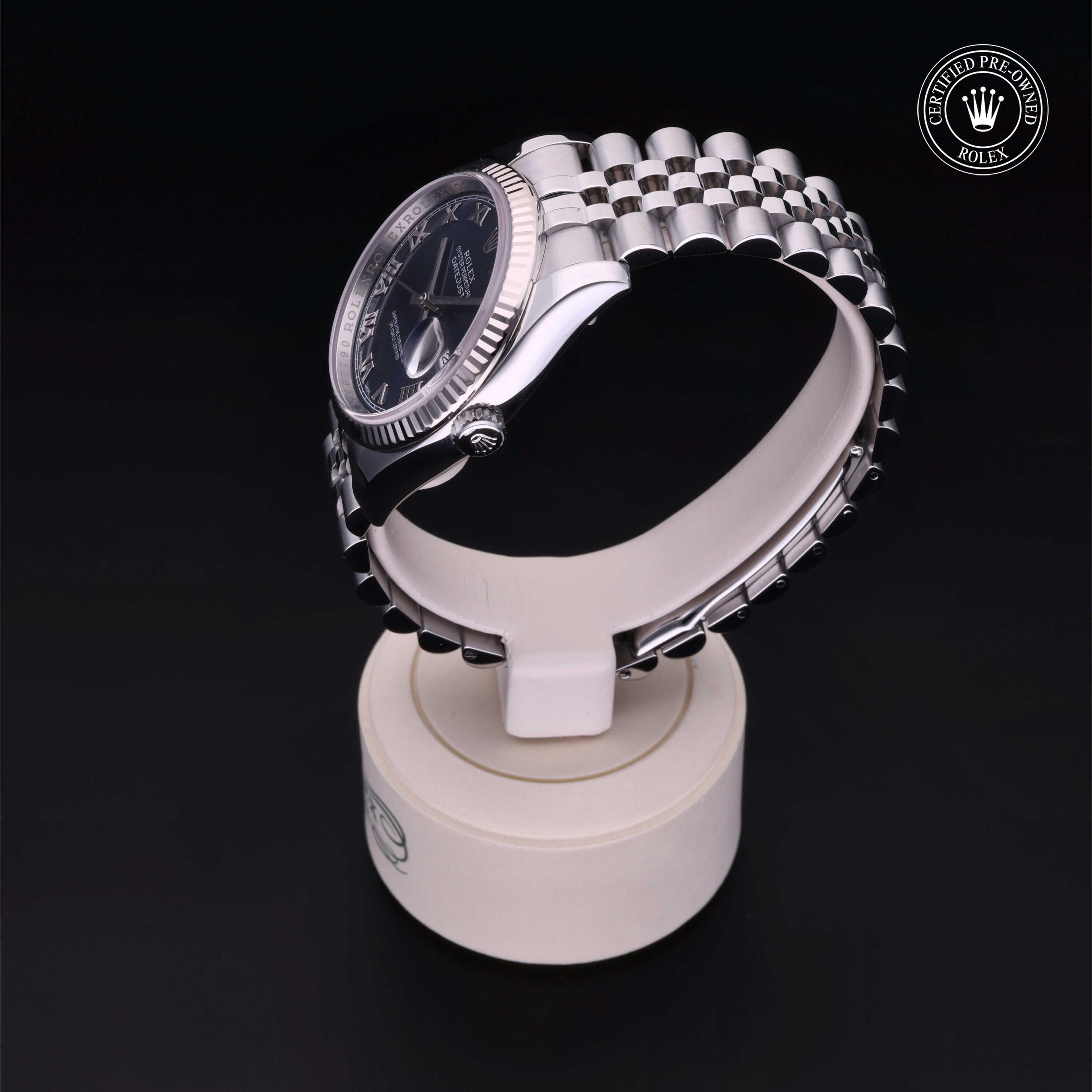 Side view of a luxury stainless steel Rolex watch with a black dial and jubilee bracelet on a white stand, featuring a certified pre-owned Rolex logo in the corner.