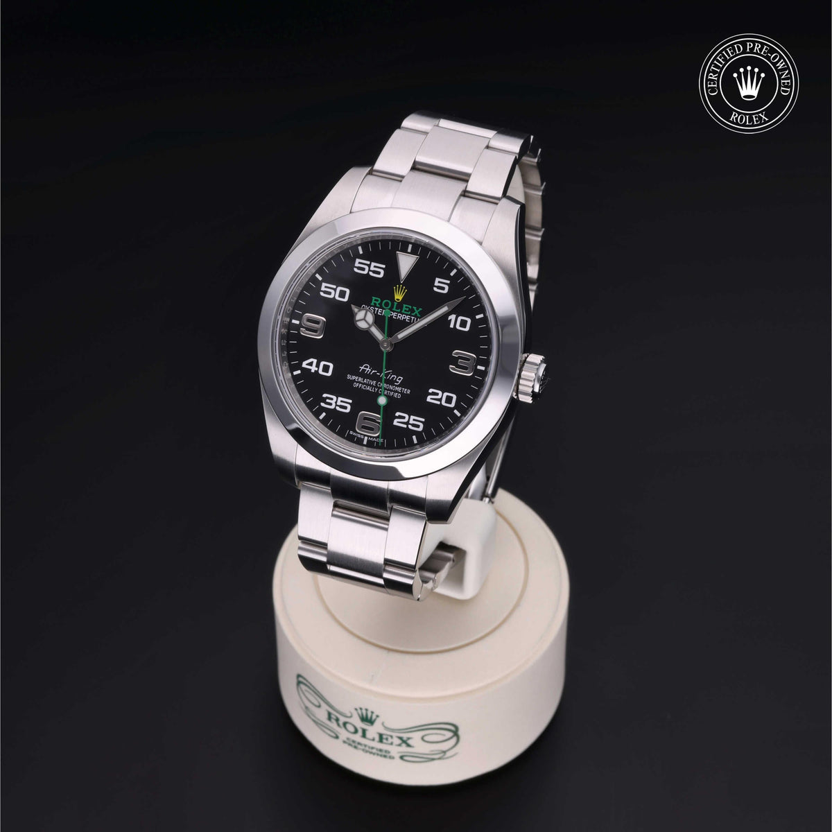 A Rolex luxury watch with a stainless steel band on a white display stand against a black background, featuring a certified pre-owned Rolex logo.