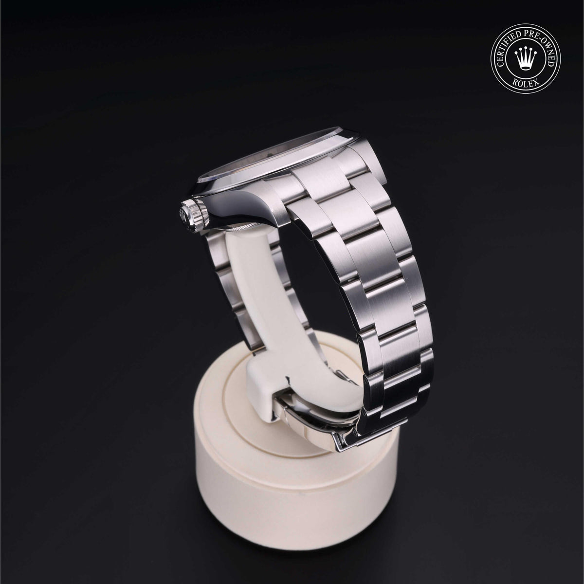 Side view of a luxury stainless steel Rolex watch with a polished bracelet displayed on a white stand against a black background, featuring a certified pre-owned Rolex logo.