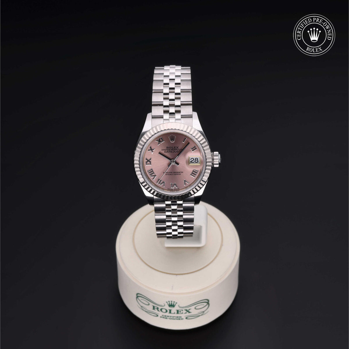Rolex luxury watch with silver metal bracelet and pink dial featuring Roman numerals, displayed on a white stand with Rolex branding against a black background.