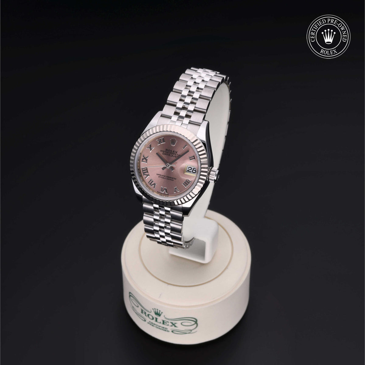 Rolex luxury watch with a silver metal band and pink dial featuring Roman numerals, displayed on a white stand against a black background with a certified pre-owned Rolex logo.