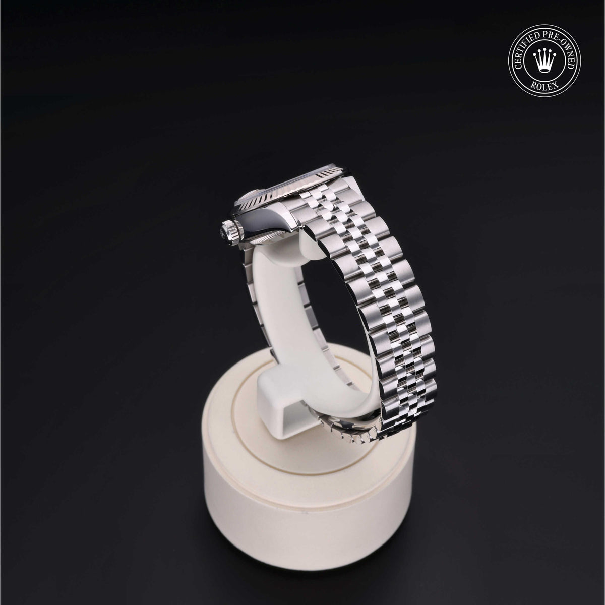 Silver-tone metal watch band with a polished finish displayed on a round cream-colored stand against a black background, featuring a Rolex Certified Pre-Owned logo.
