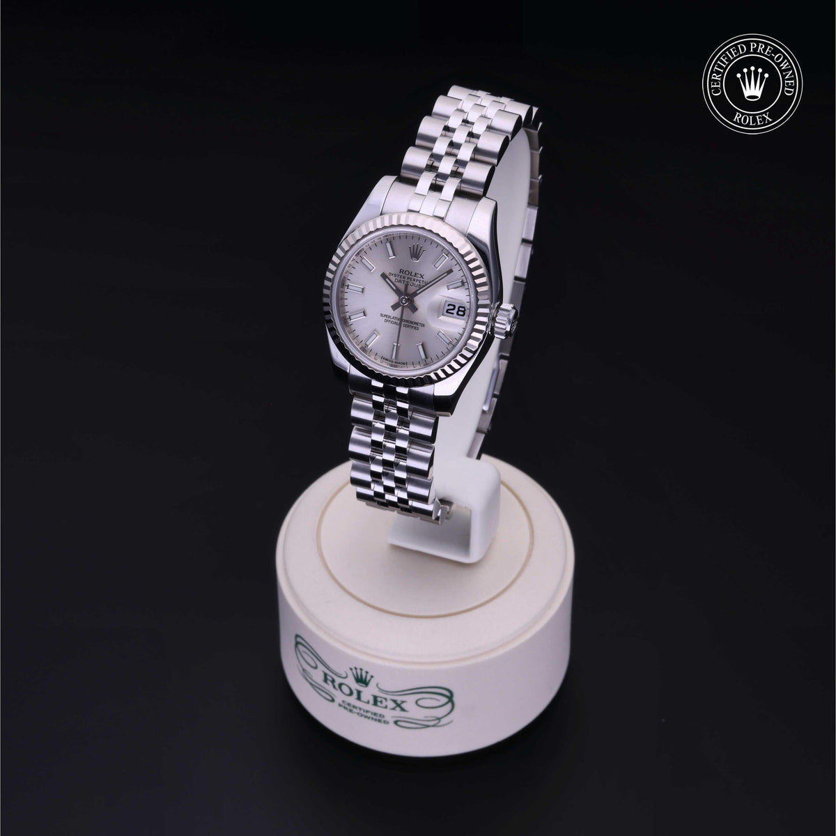 Silver Rolex wristwatch with fluted bezel and date display on a white round stand, featuring a five-link Jubilee bracelet and a 