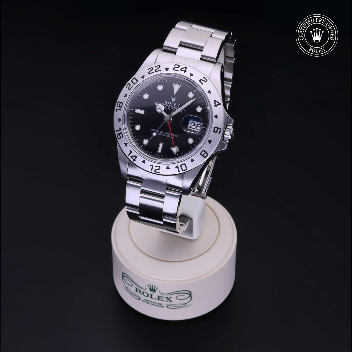 Close-up of a stainless steel Rolex luxury watch with a black dial and date display on a branded stand, featuring a certified pre-owned Rolex logo in the corner.