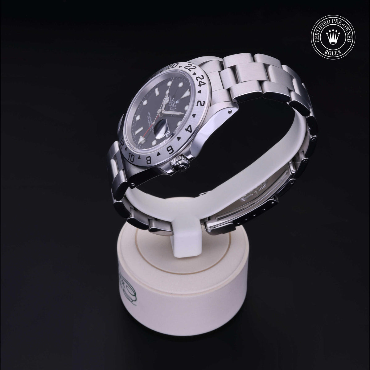 Close-up of a luxury stainless steel Rolex watch with a black dial and a 24-hour bezel, displayed on a white stand against a black background.
