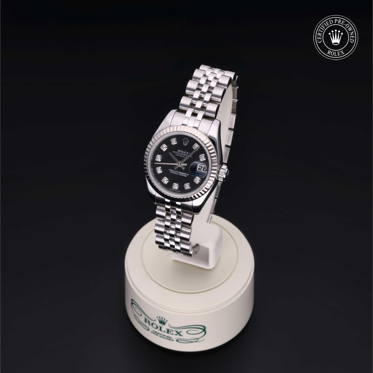 Luxury silver Rolex watch with black dial and diamond hour markers displayed on a white stand against a black background with a certified pre-owned logo.