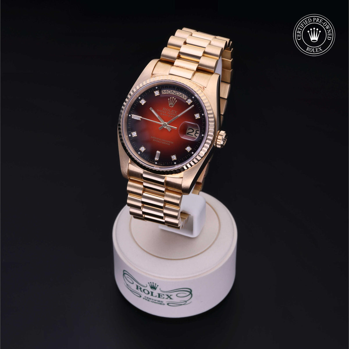 Luxury Rolex watch with a gold bracelet and gradient red to black dial featuring diamond hour markers, displayed on a branded stand with a certified pre-owned emblem.