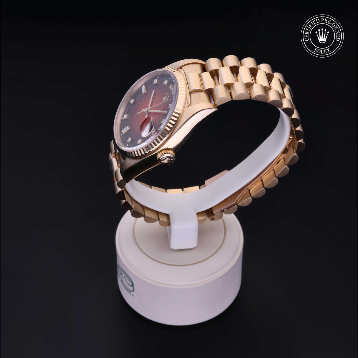 Gold-toned luxury wristwatch with a polished bracelet and fluted bezel displayed on a white stand against a black background, featuring a certified pre-owned Rolex emblem.