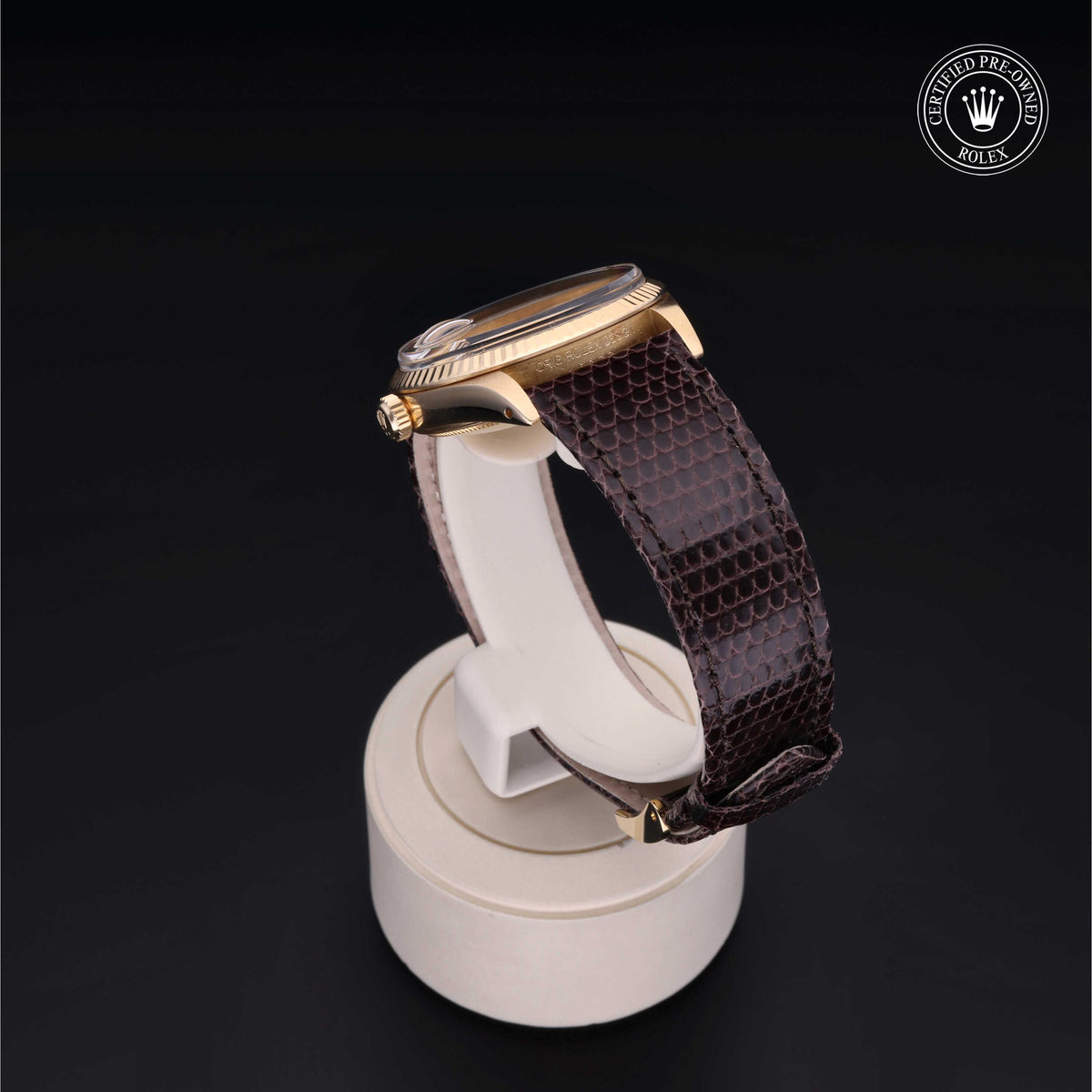 Side view of a luxury wristwatch with a gold case and textured brown leather strap displayed on a white stand against a black background, featuring a Certified Pre-Owned Rolex emblem.