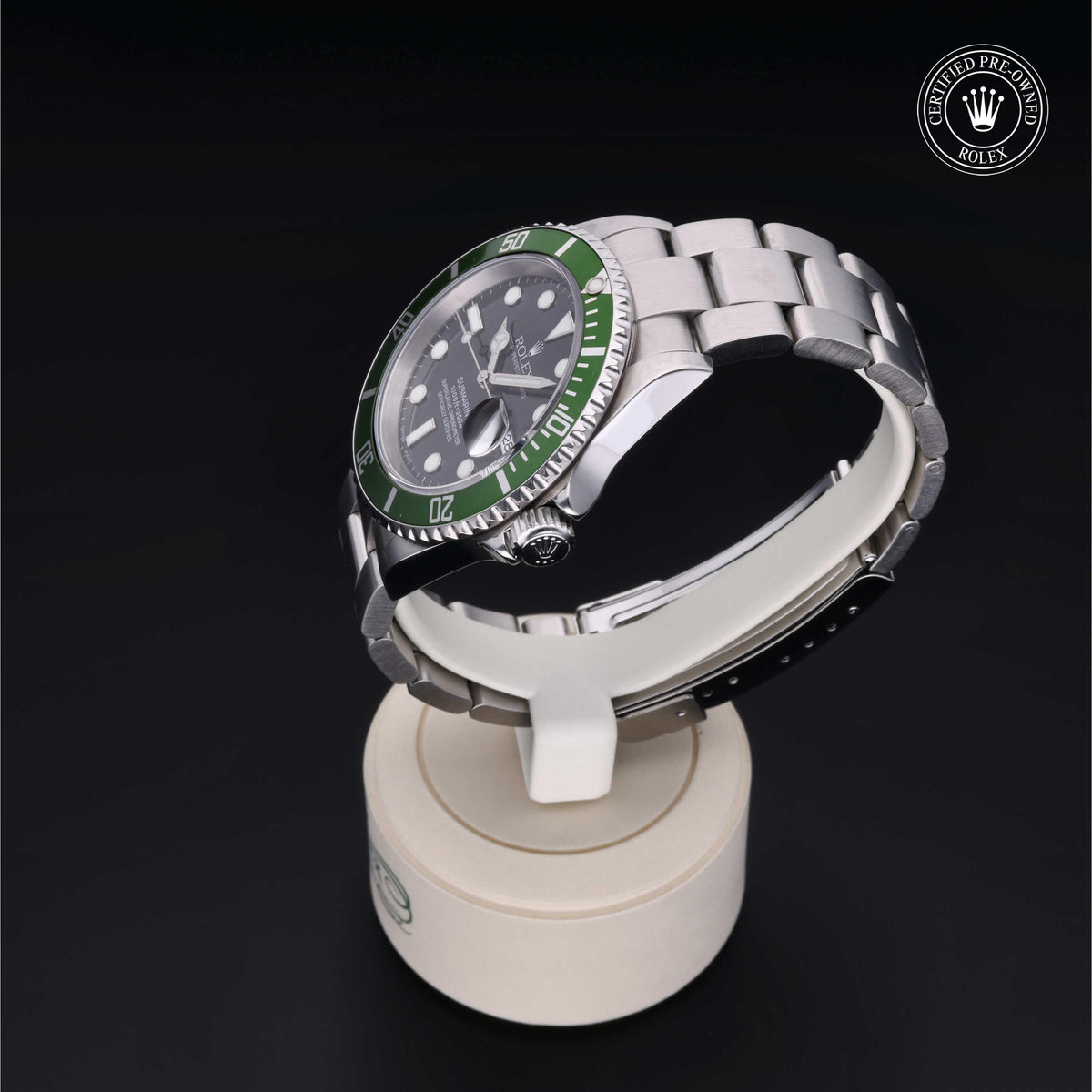 Rolex stainless steel luxury watch with green bezel and black dial, displayed on a white stand against a black background, featuring a certified pre-owned Rolex logo.
