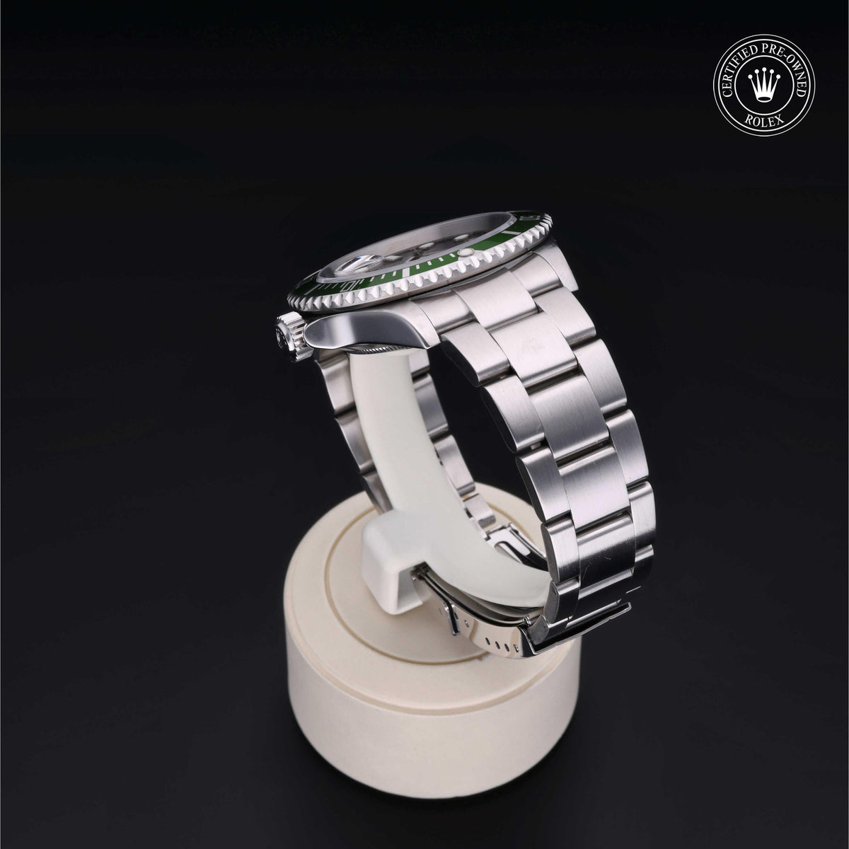 Side view of a silver stainless steel Rolex watch with a green bezel displayed on a white stand against a black background, with a certified pre-owned Rolex logo in the corner.
