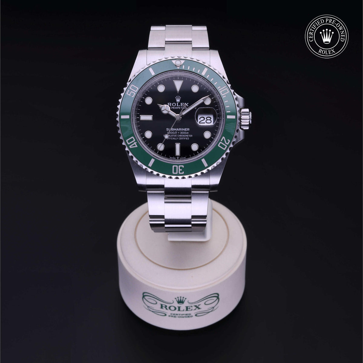 A Rolex Submariner watch with a green bezel and black dial displayed on a branded Rolex stand against a black background, featuring a certified pre-owned logo in the corner.