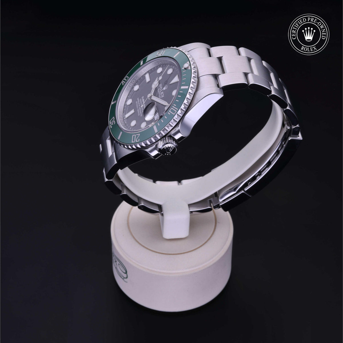 Close-up of a luxury Rolex watch with a green bezel displayed on a white stand against a black background, showing its stainless steel bracelet and detailed dial.
