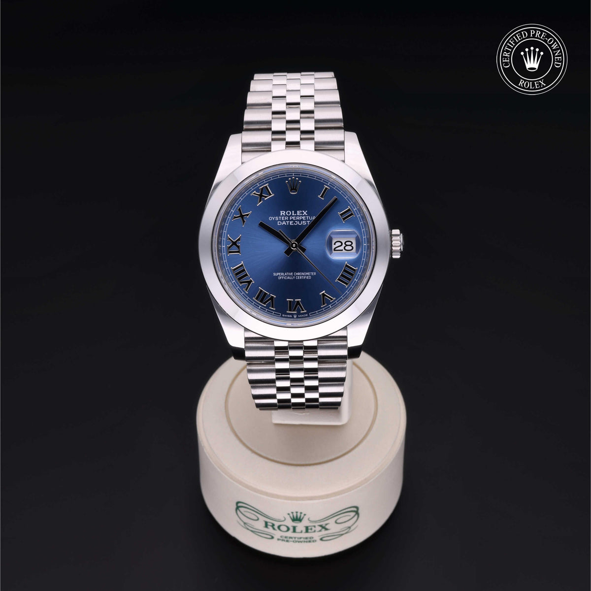 Rolex wristwatch with a blue dial and Roman numeral markers displayed on a round white stand with Rolex branding against a dark background, featuring a certified pre-owned logo.